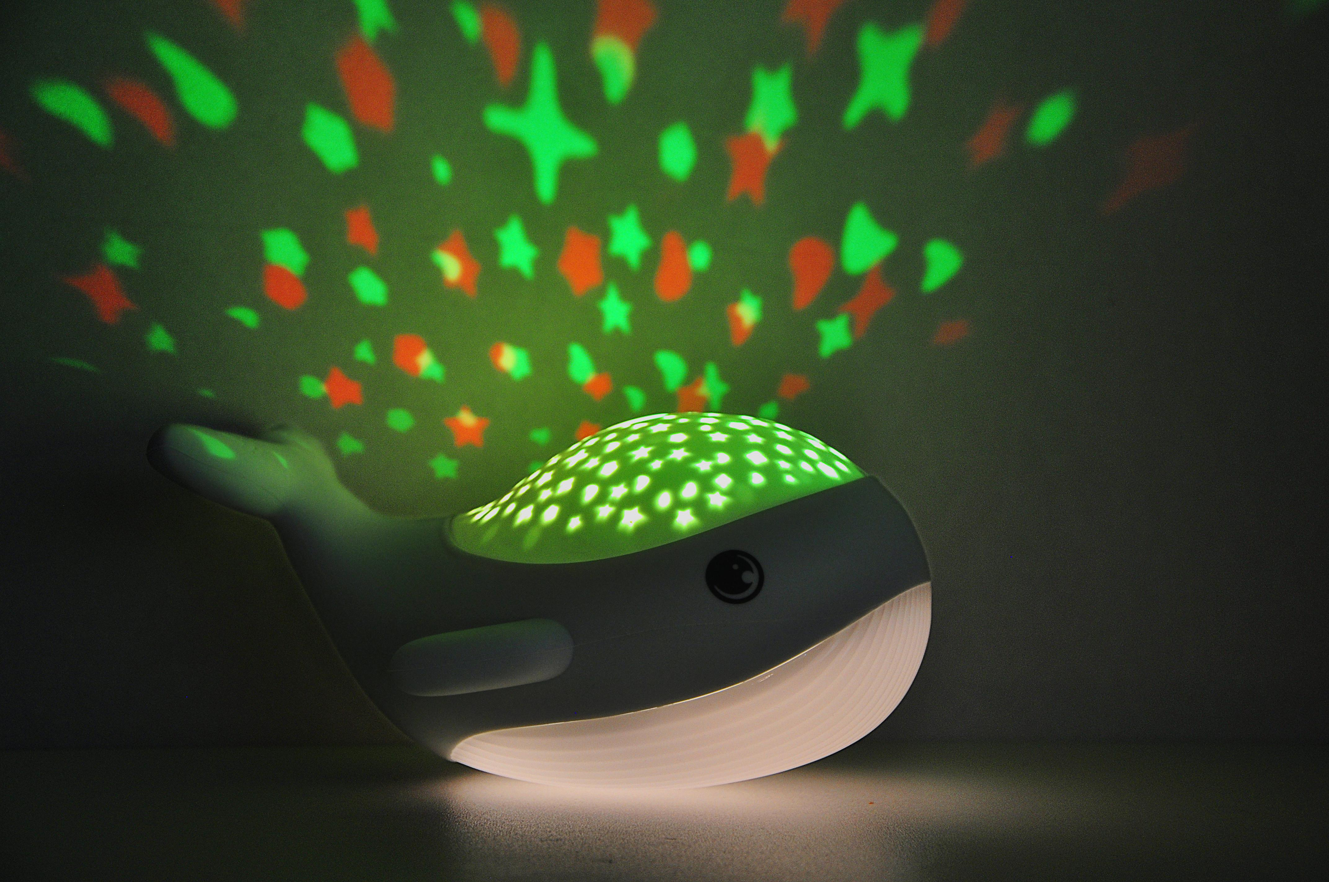 Ein walförmiges Nachtlicht, das Sterne projiziert. Grüne und orangefarbene Sterne an einer Wand.