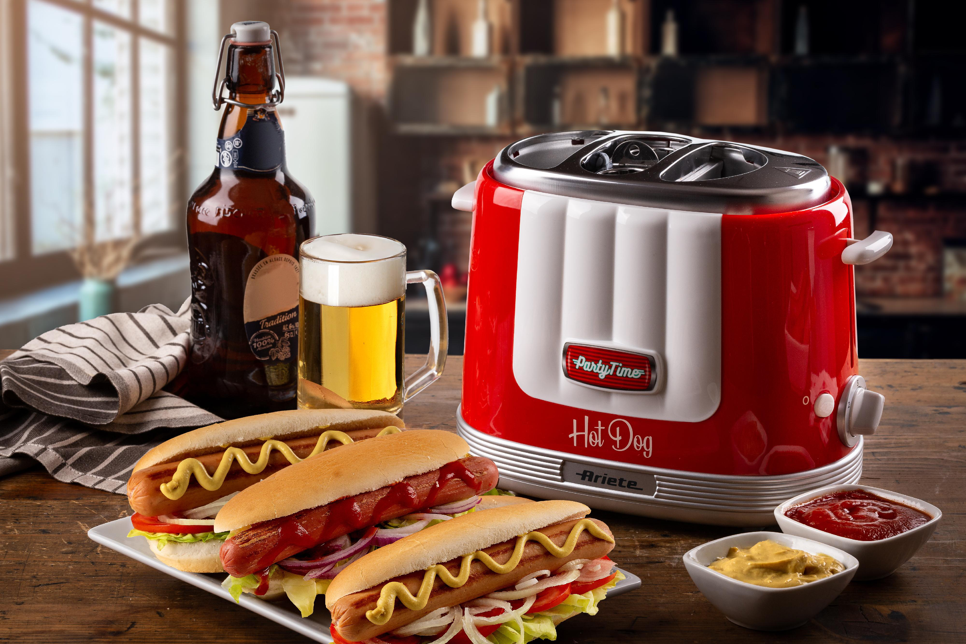 Ein roter Hotdog-Maker mit Hotdogs und Gewürzen auf einem Tisch. Bier und ein Bierkrug im Hintergrund.