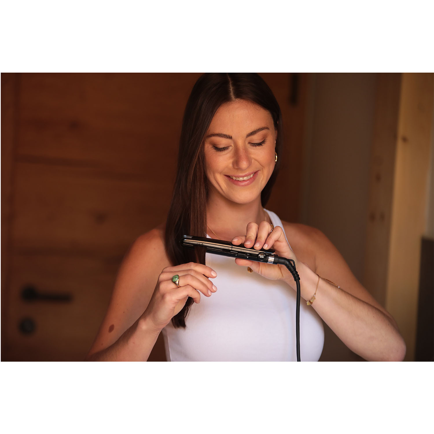 Donna sorridente, tiene una piastra per capelli nera. Indossa una canotta bianca.