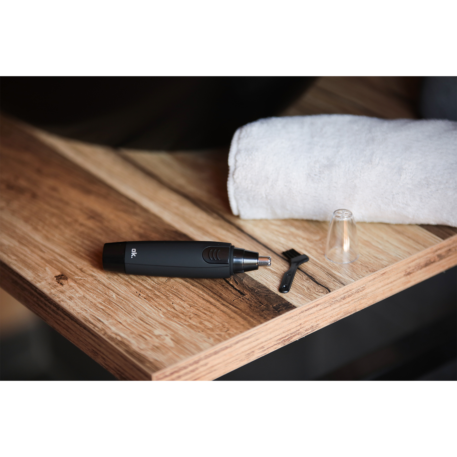 Tagliacapelli nasali nero e accessori su una superficie in legno, asciugamano bianco sullo sfondo.
