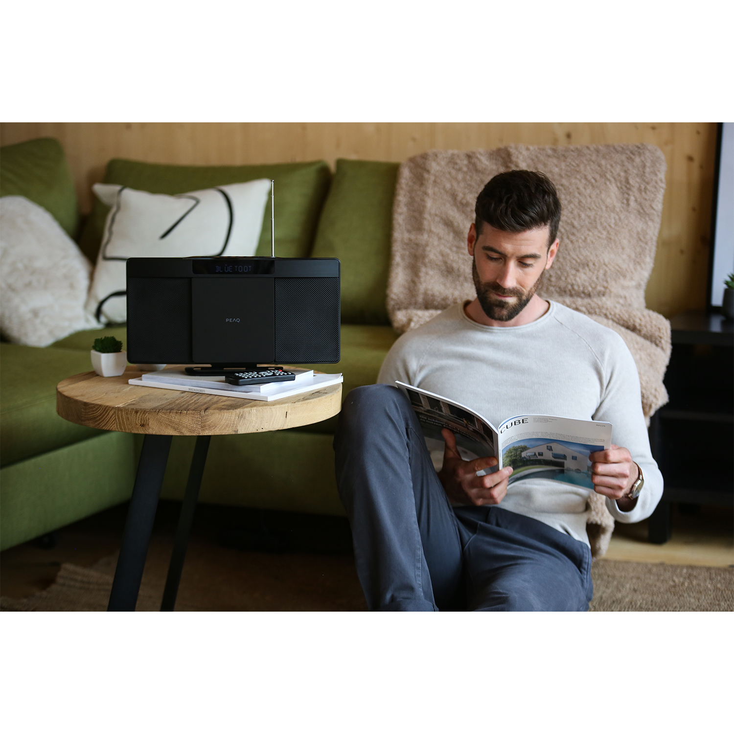 Uomo legge una rivista vicino a uno stereo e a un divano. Un tavolino contiene una pianta e un telecomando.