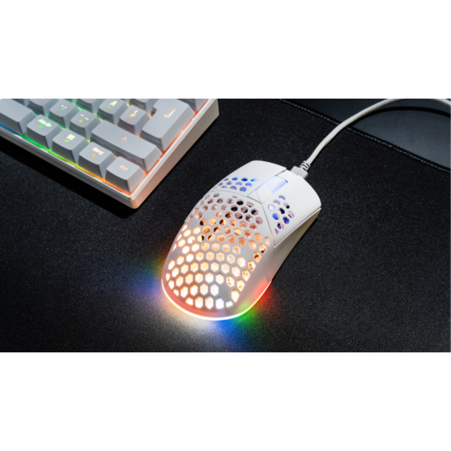 White gaming mouse with honeycomb design, lit with rainbow colors, near a keyboard on black surface.