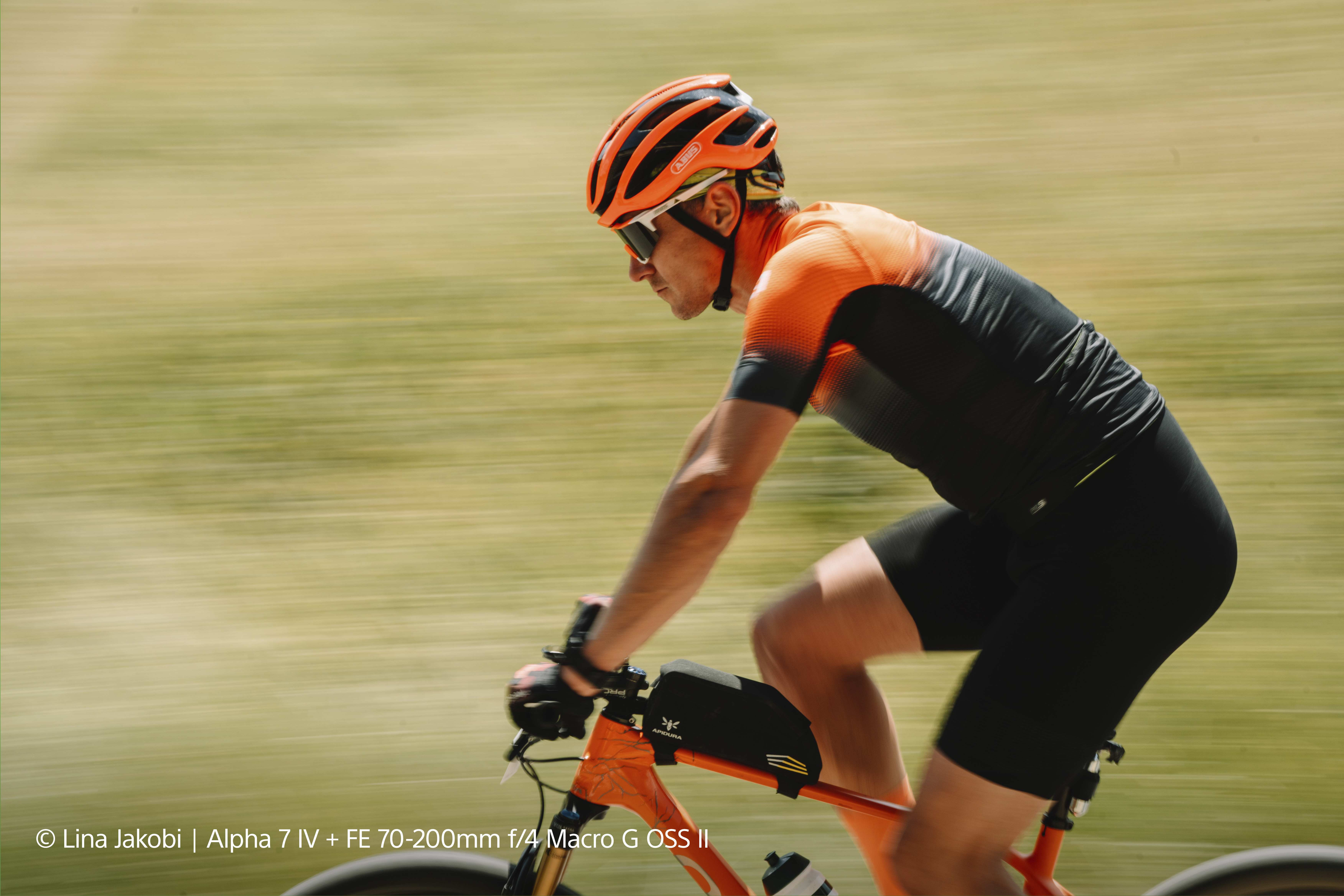 Radfahrer mit orangefarbenem Helm und Trikot, unscharfer Hintergrund.