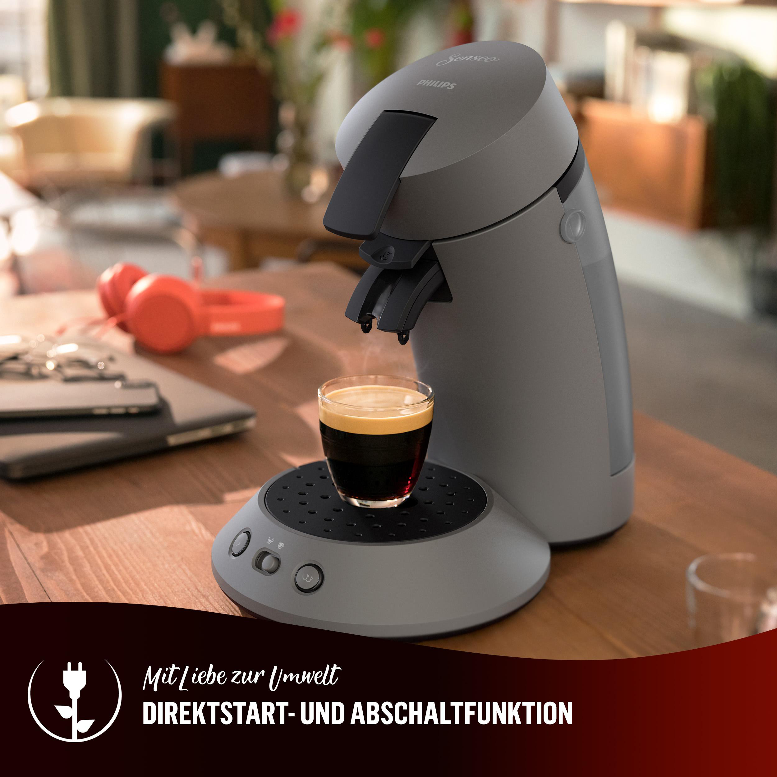 Eine graue Kaffeemaschine mit einer Tasse Kaffee und Text auf Deutsch auf einem Holztisch.