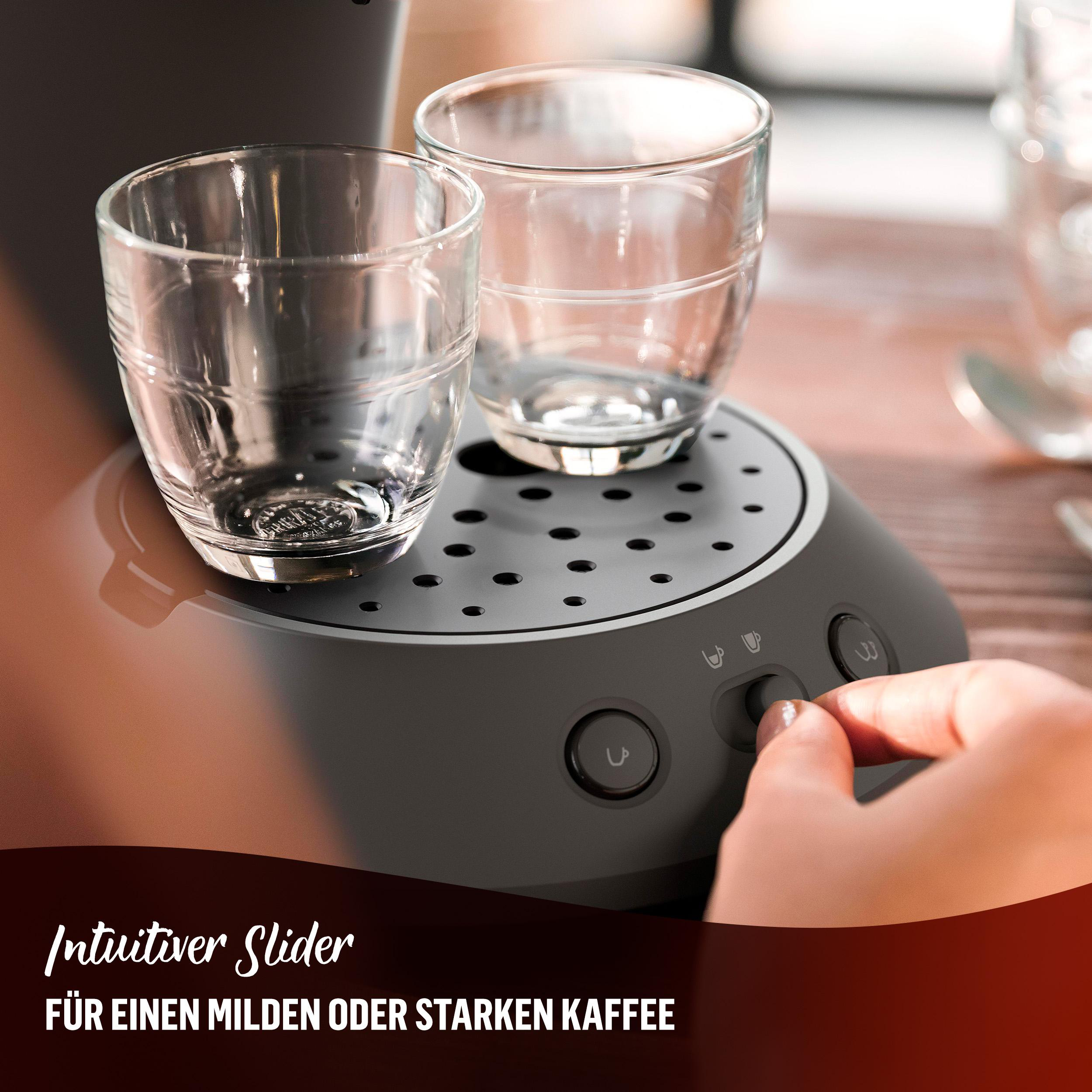 Nahaufnahme einer Kaffeemaschine mit zwei leeren Gläsern, eine Hand berührt einen Knopf.