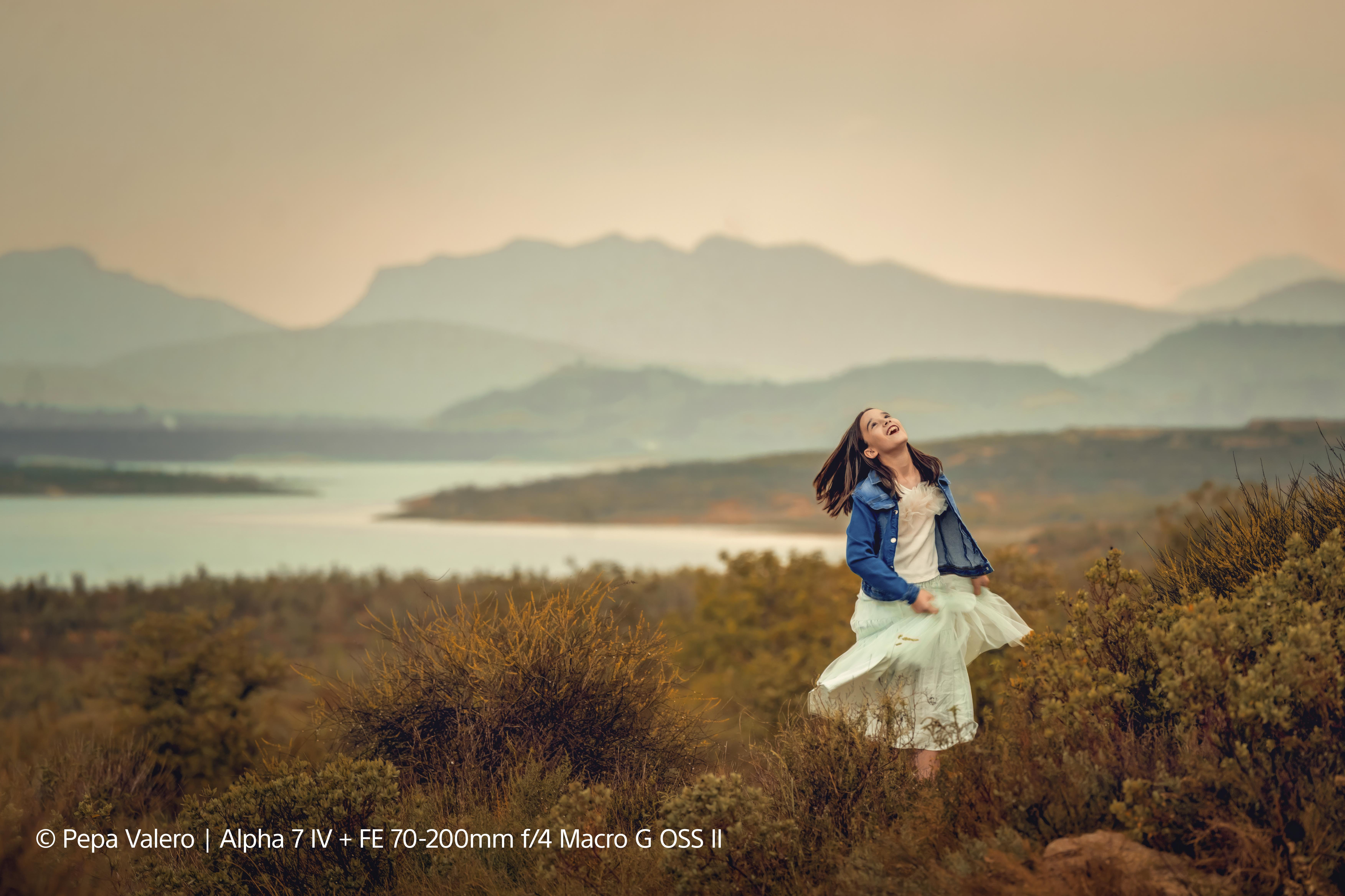 Mädchen mit weißem Kleid lacht auf einem Feld. Berge und See im Hintergrund.