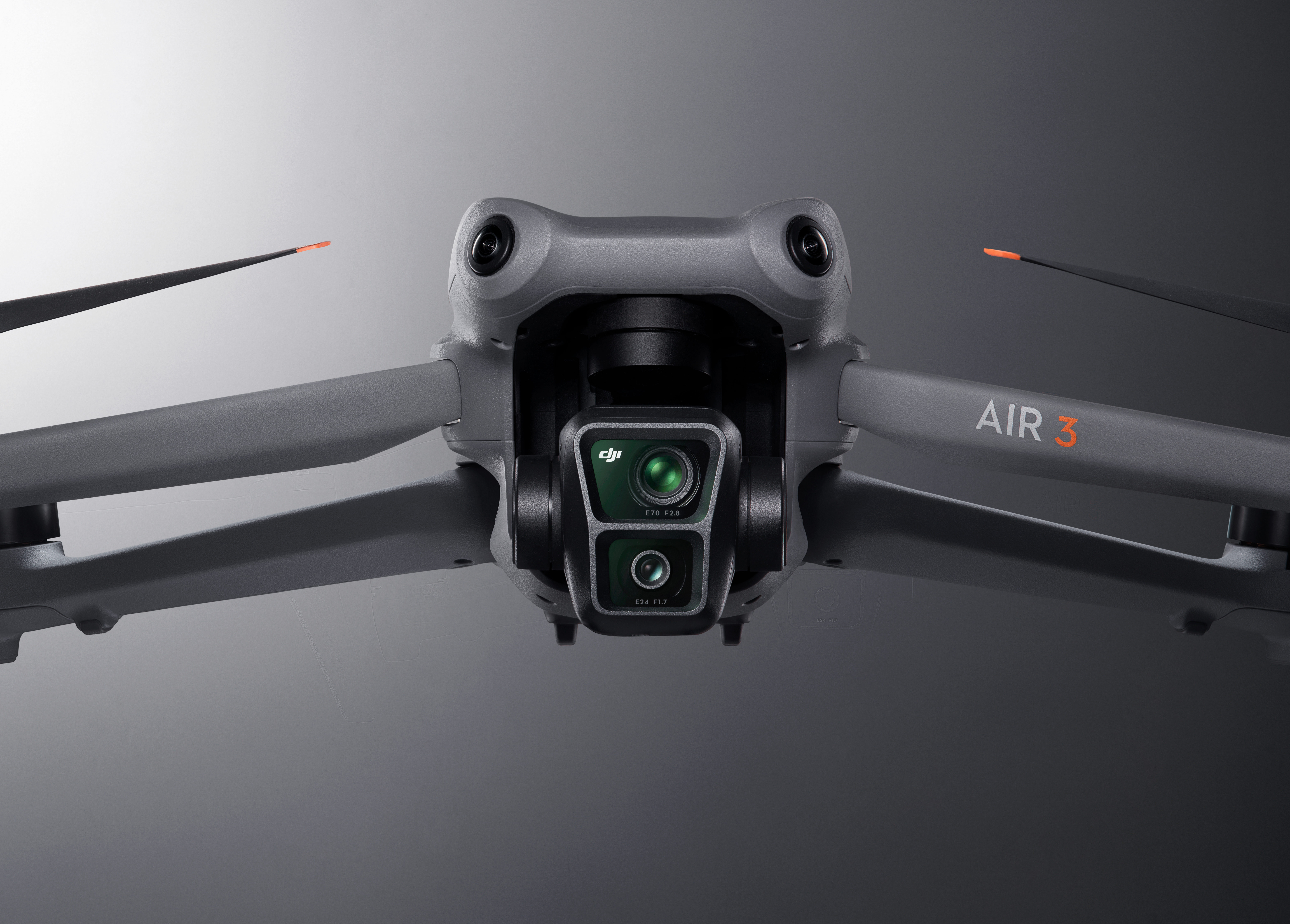 Un drone grigio DJI Air 3 centrato. La sua fotocamera è a fuoco, con eliche che si estendono. Lo sfondo è grigio.