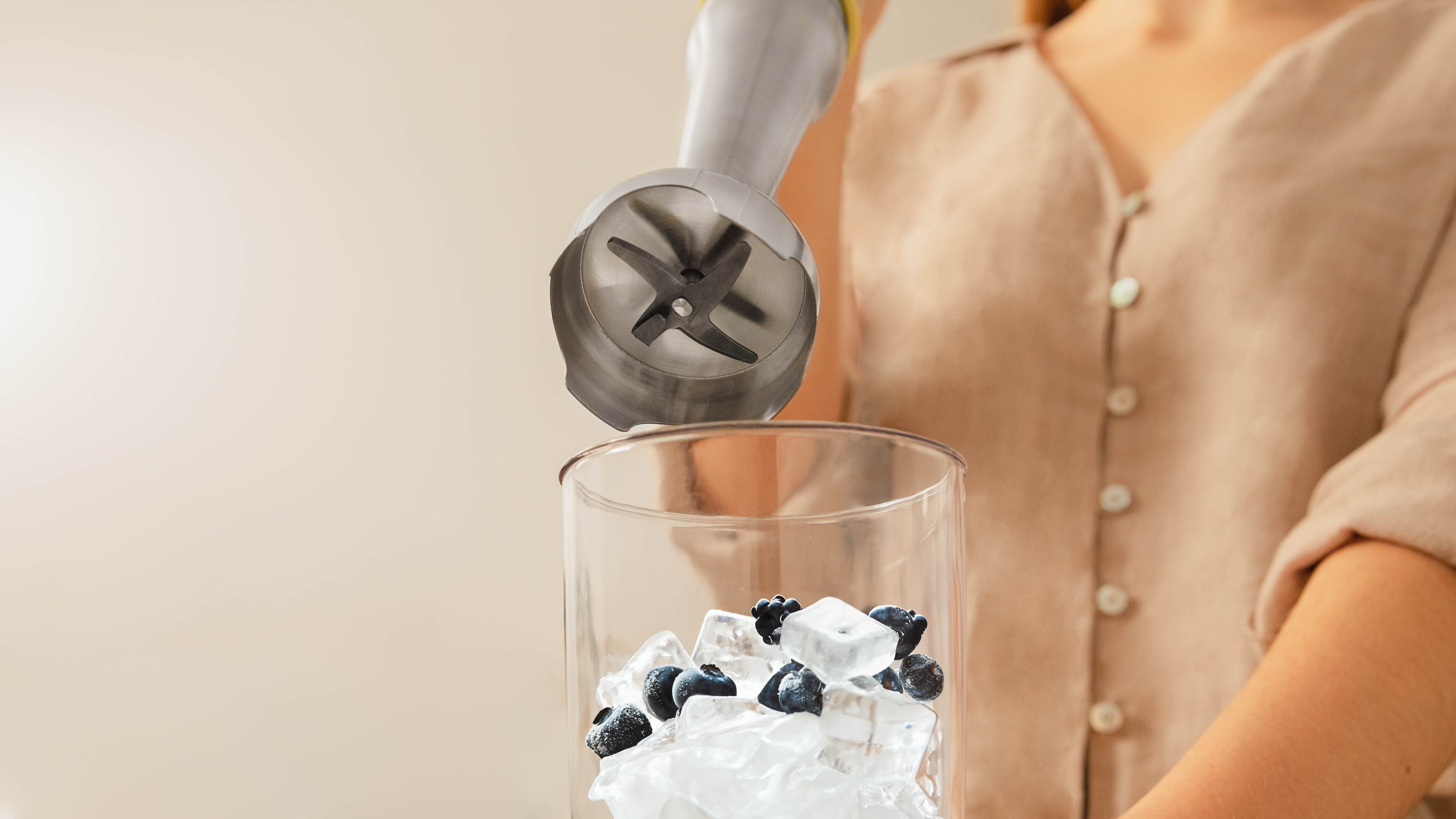 Una persona usando una batidora de mano para mezclar cubitos de hielo y arándanos en un vaso alto.