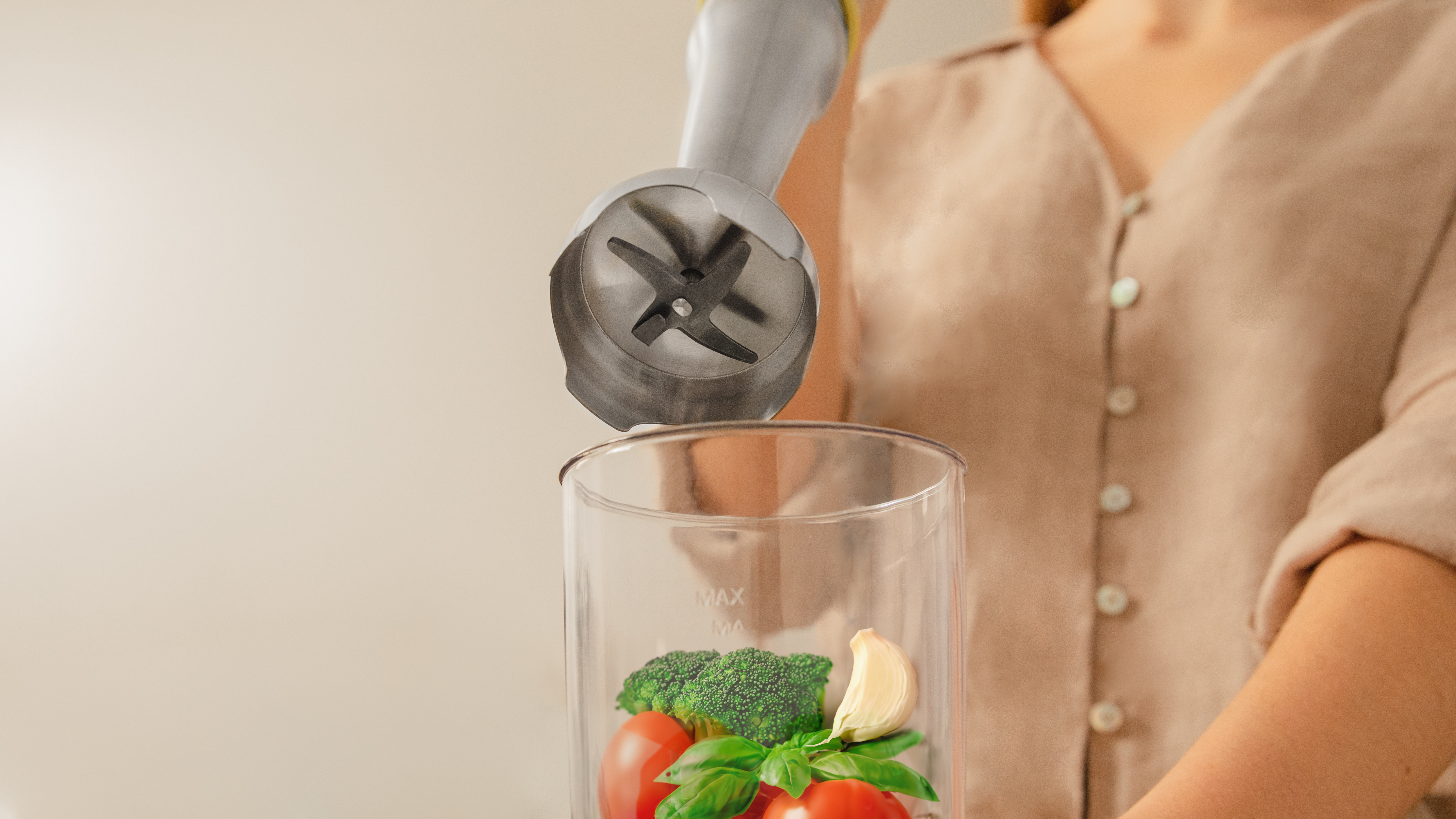 Una persona mezclando ingredientes con una batidora de inmersión, tomates, brócoli y ajo son visibles.
