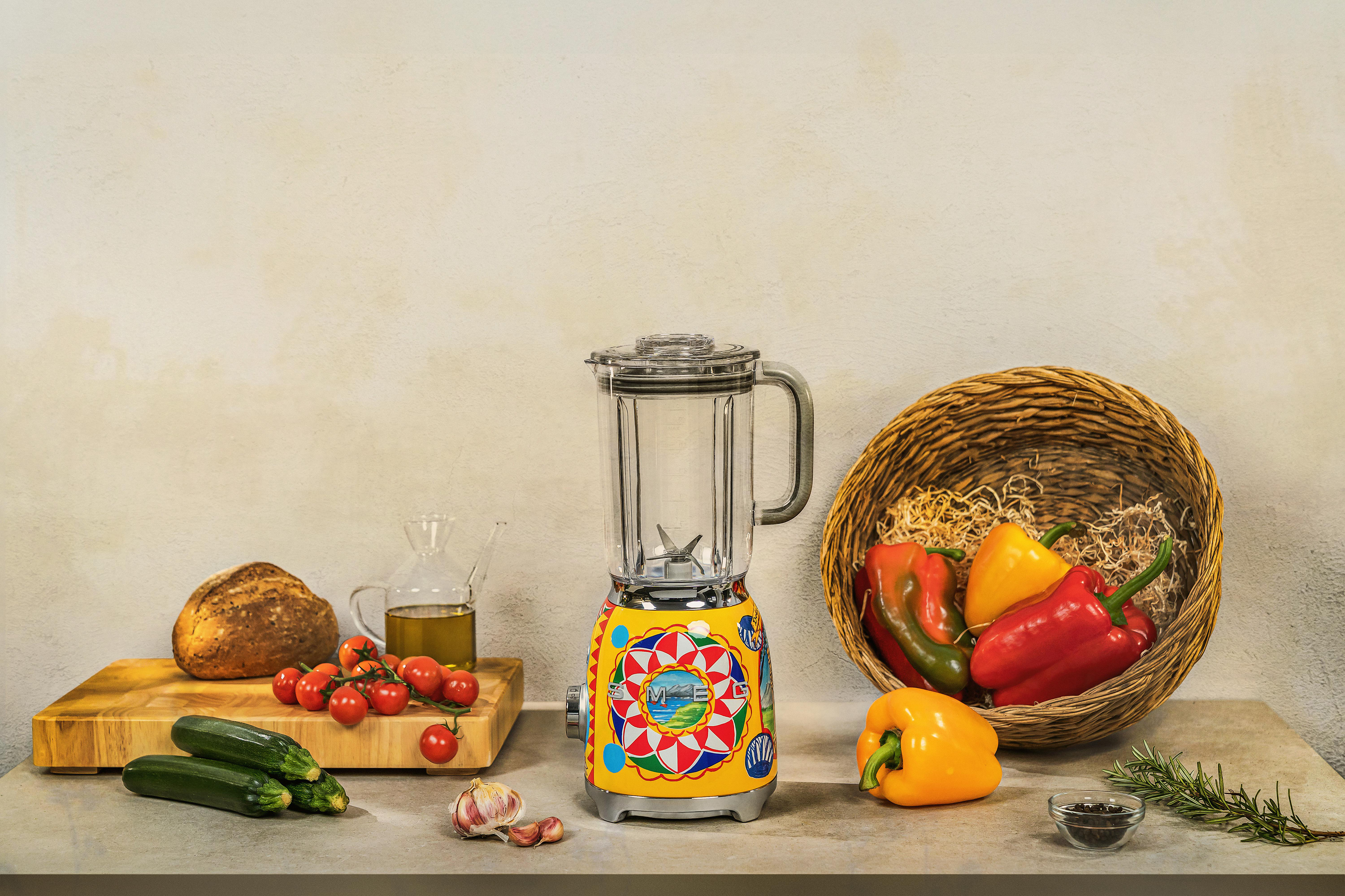 Ein verzierter Mixer steht auf einem Tisch mit Gemüse, Brot und Olivenöl.