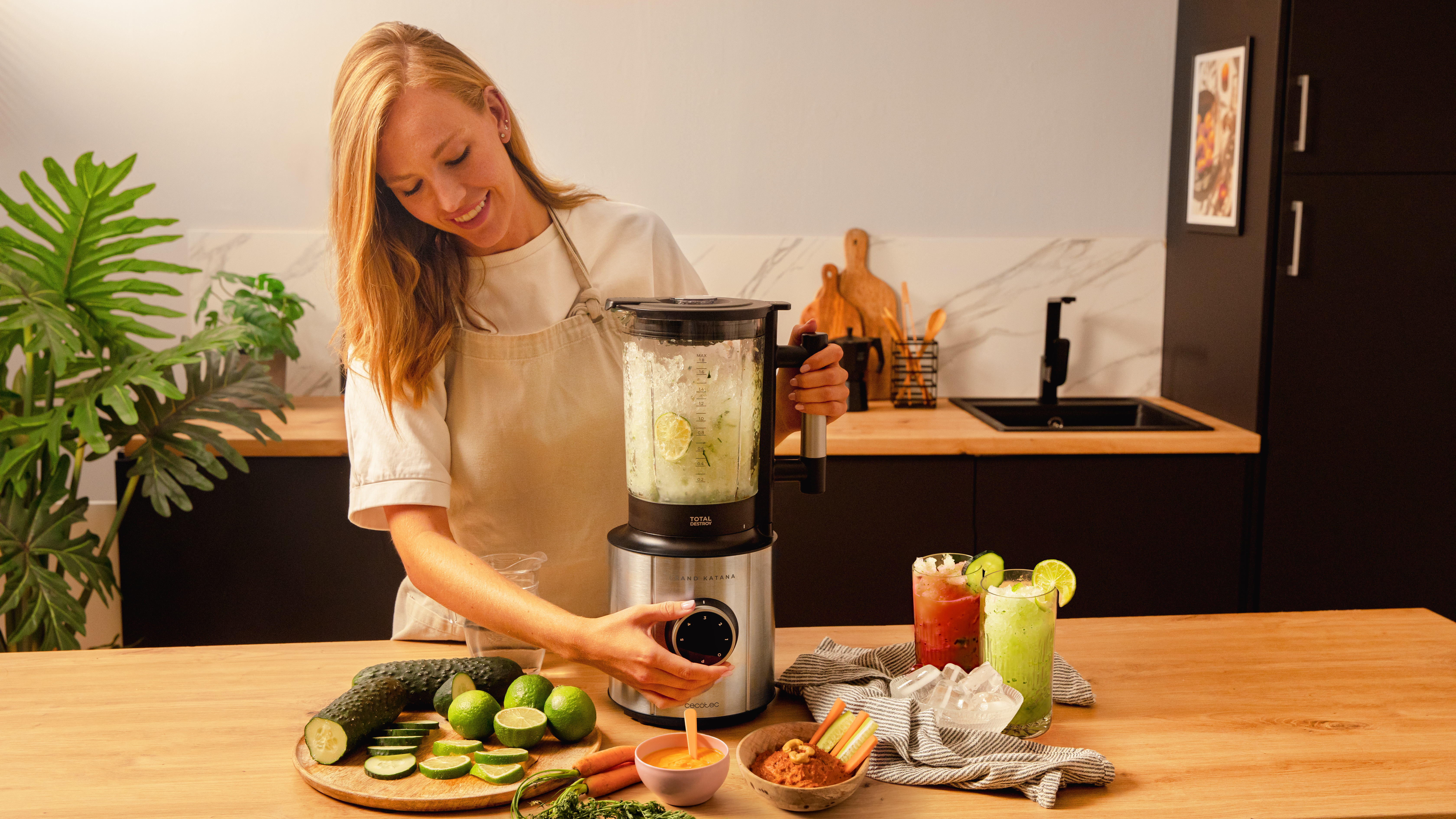 Mujer rubia preparando un batido. Licuadora, ingredientes y batidos en mesa de madera. Fondo de cocina.