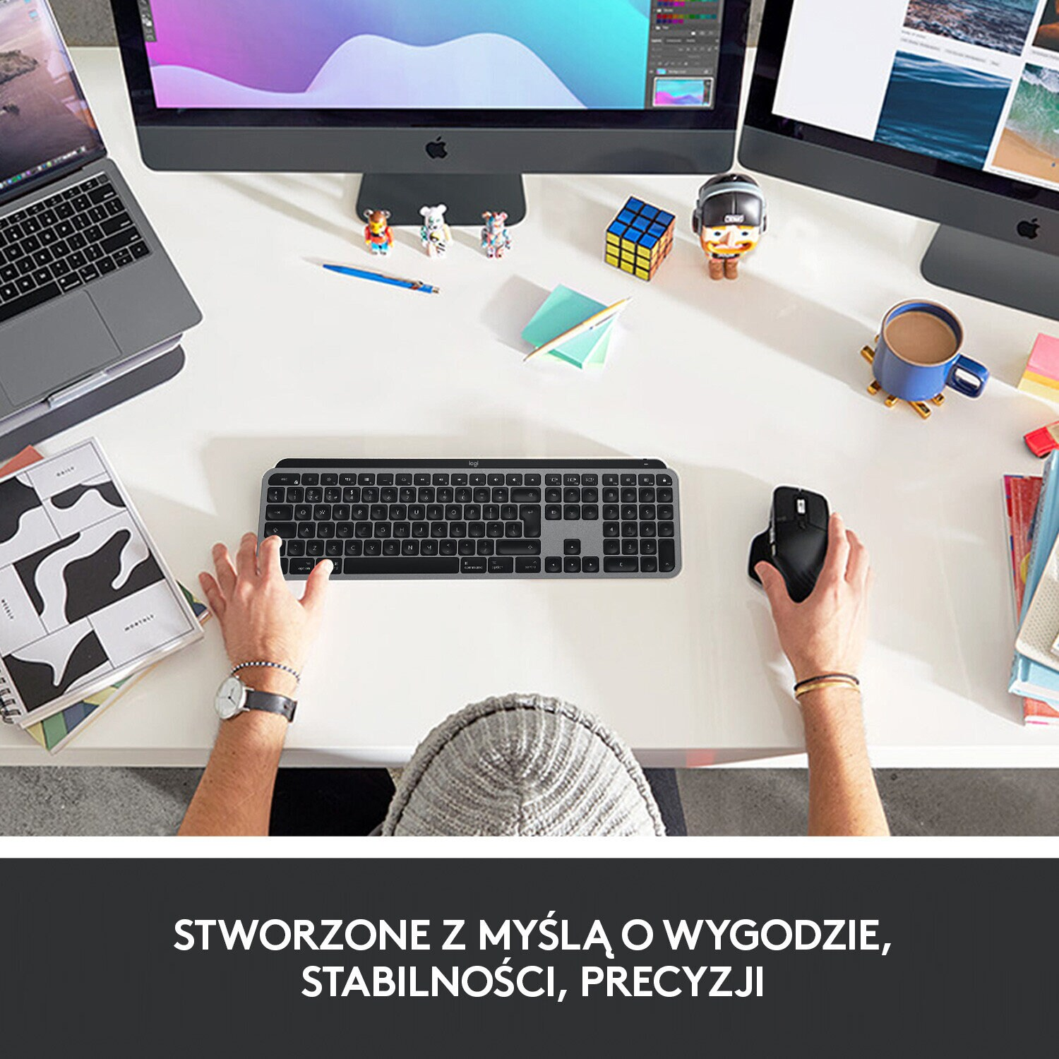 Osoba przy biurku z klawiaturą, myszą i monitorami. Widok z góry. Tekst na dole.
