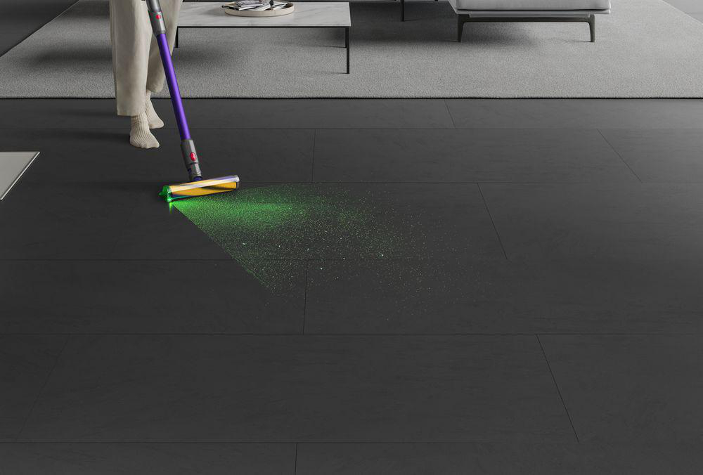 Ein Dyson-Staubsauger mit grünem Laserlicht reinigt einen dunklen Boden. Eine Person benutzt ihn.