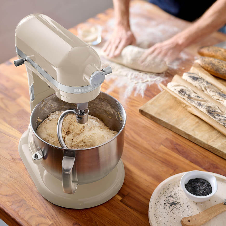 Egy bézs színű KitchenAid robotgép tésztát kever egy rozsdamentes acél tálban. Egy másik személy dolgozik.