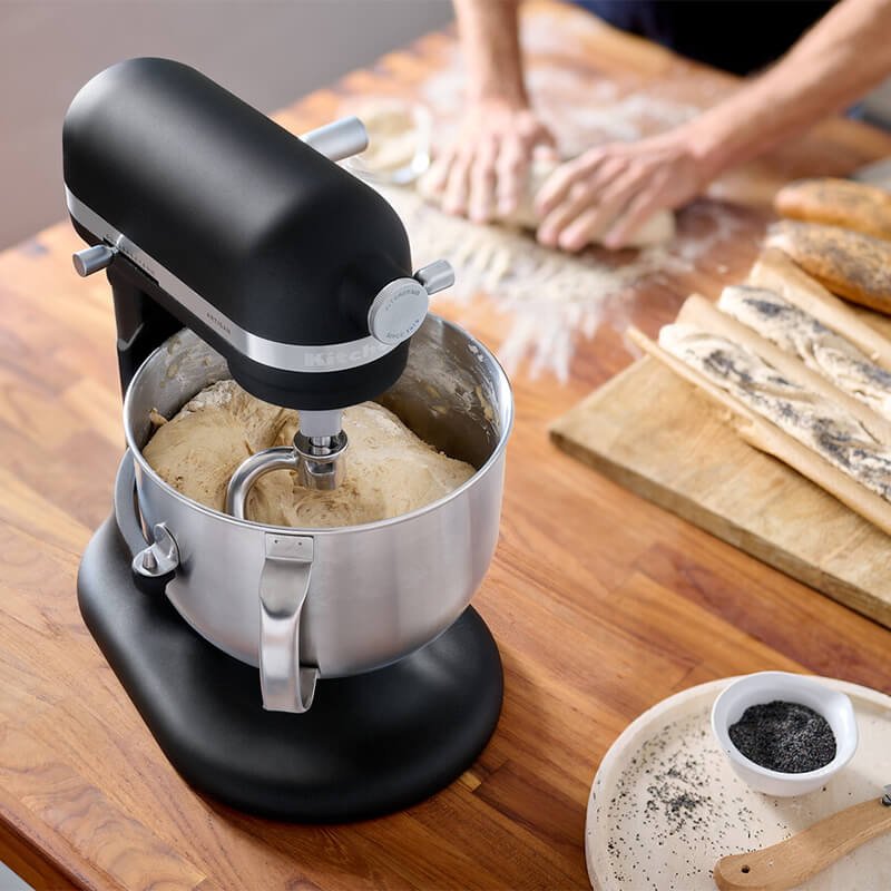 Fekete KitchenAid robotgép tésztával a tálban. Fa asztal, mák és személy a háttérben.
