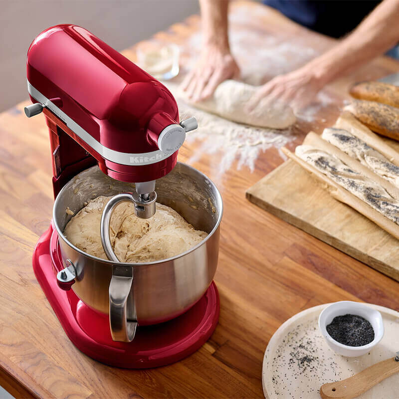 Piros állványos mixer tésztát kever, személy lisztet szitál.