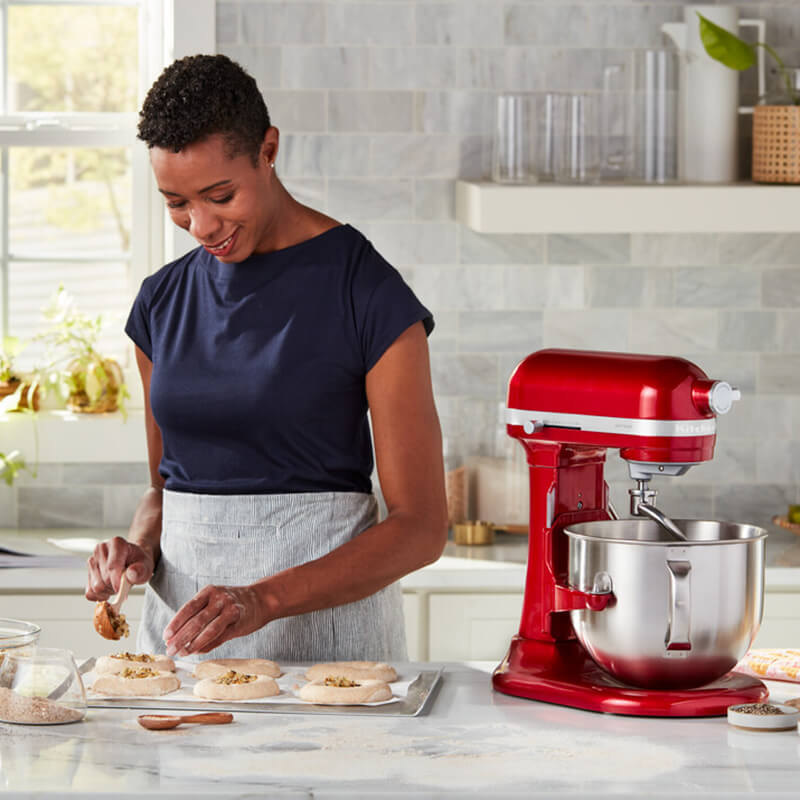 Egy nő ételt készít, tölteléket helyez a péksüteményekre, jobbra egy piros KitchenAid keverővel.