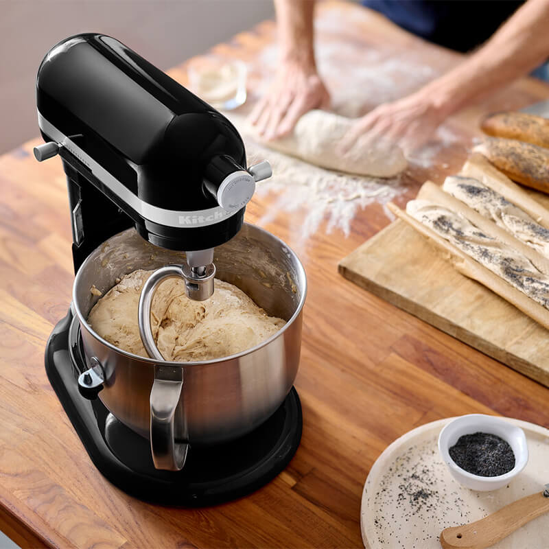 Fekete KitchenAid mixer tésztát kever tálban, fa asztal, összetevők a háttérben.