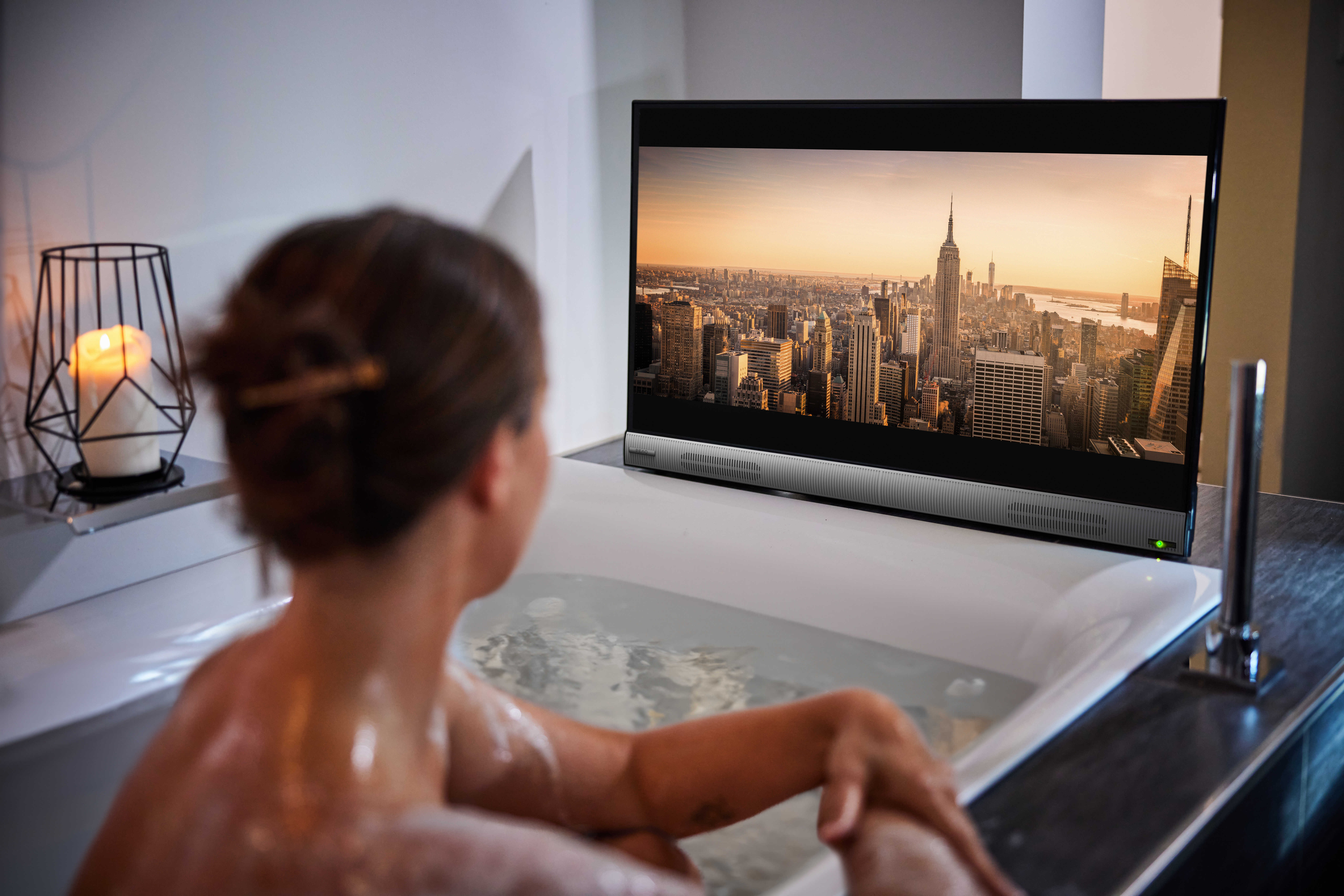 Frau in der Badewanne, die fernsieht. Der Fernseher ist auf einem Ständer montiert.