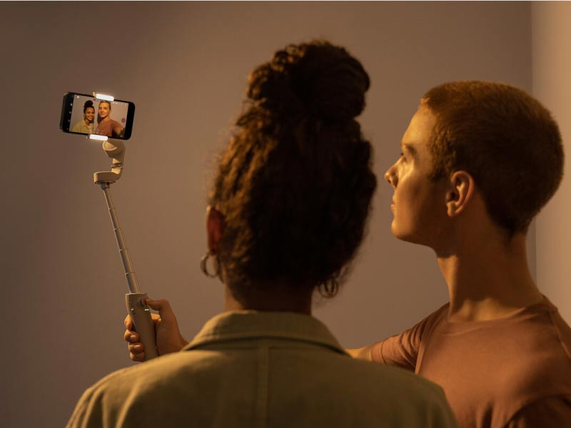 İki kişi telefon ve selfie çubuğu ile selfie çekiyor, her ikisi de halka ışıkla aydınlatılıyor.