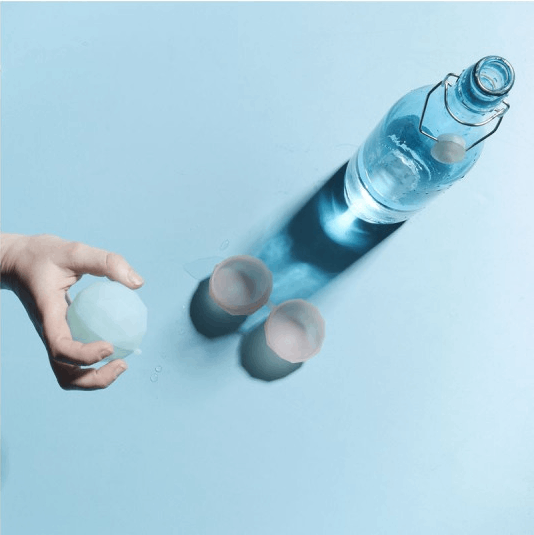 Mano sosteniendo una bola de hielo sobre dos vasos y una botella de agua. Fondo azul.