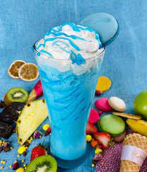Üzeri krem şanti ve mavi makaron ile süslenmiş mavi bir milkshake. İçeceği meyveler çevreliyor.