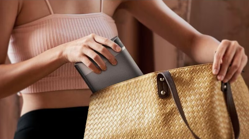 Une femme met un haut-parleur rectangulaire gris dans un sac à main beige tissé.