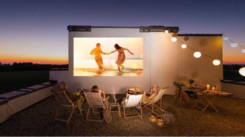 Des gens regardent un film sur un écran en plein air, avec des lumières et un coucher de soleil.