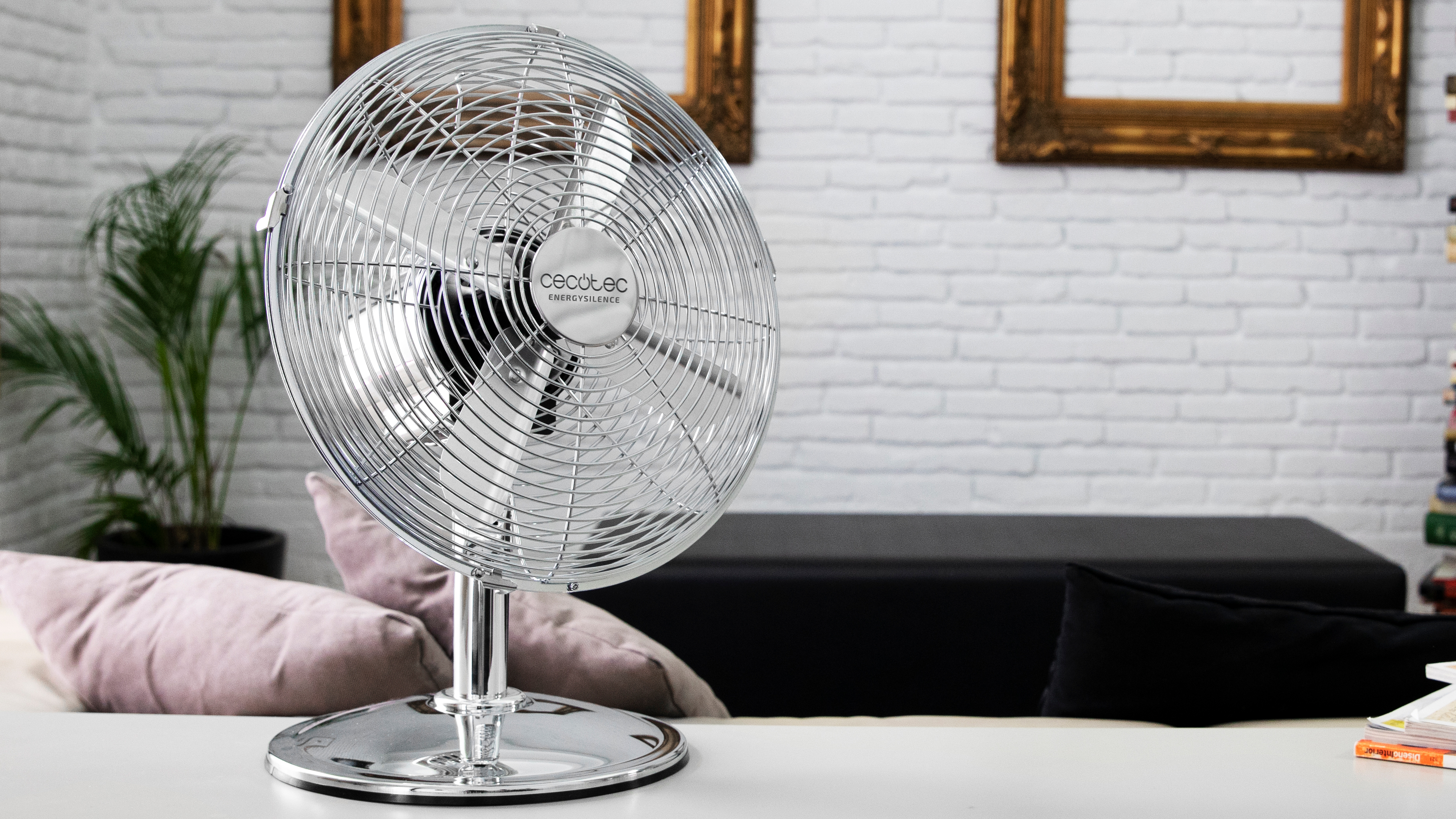 Un ventilador cromado sobre una mesa, con un fondo de pared de ladrillos blancos.
