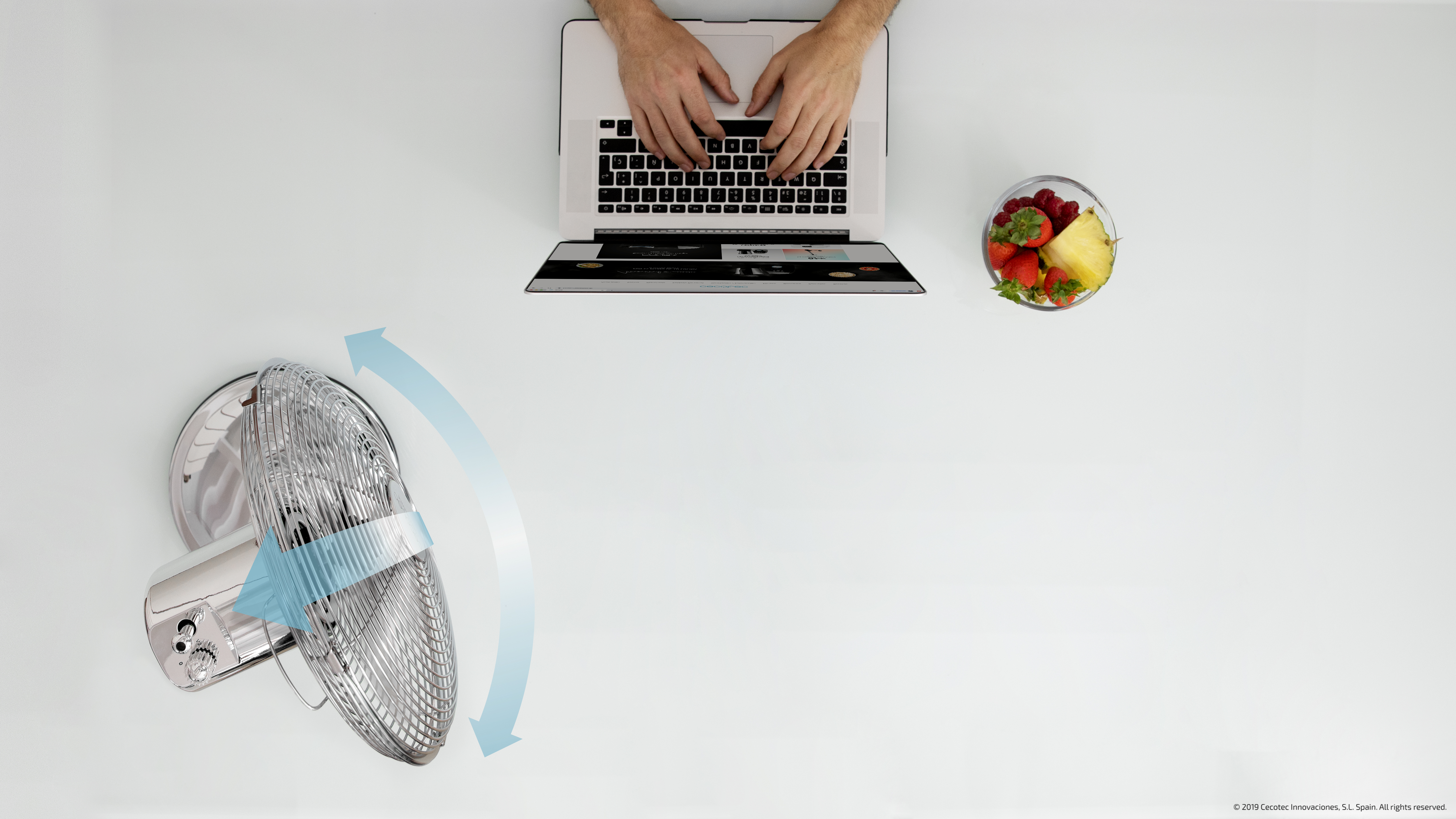 Una persona usa una laptop y un frutero sobre una mesa blanca. Un ventilador está a la izquierda.