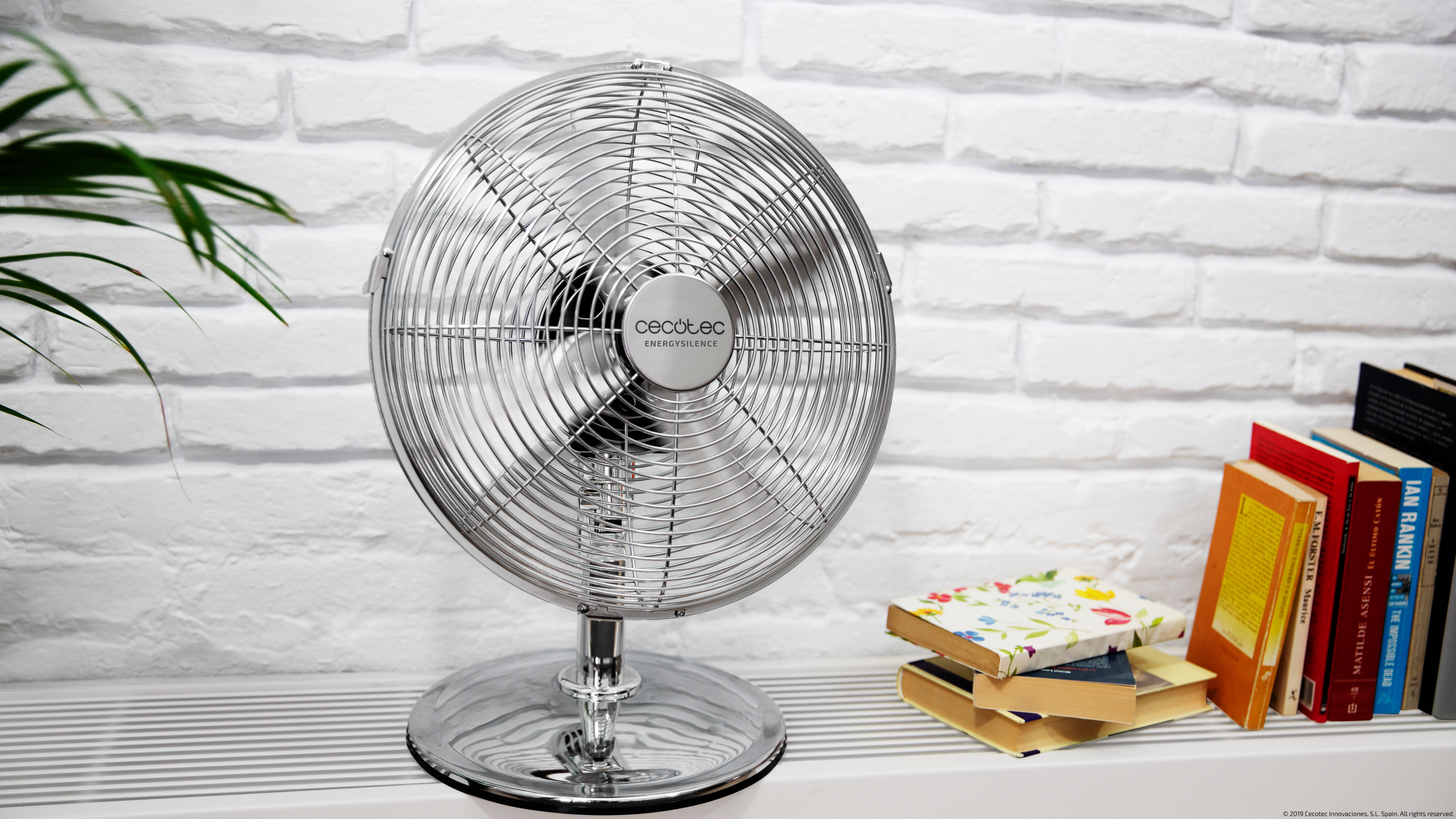 Un ventilador plateado sobre un soporte, frente a una pared de ladrillo y libros.
