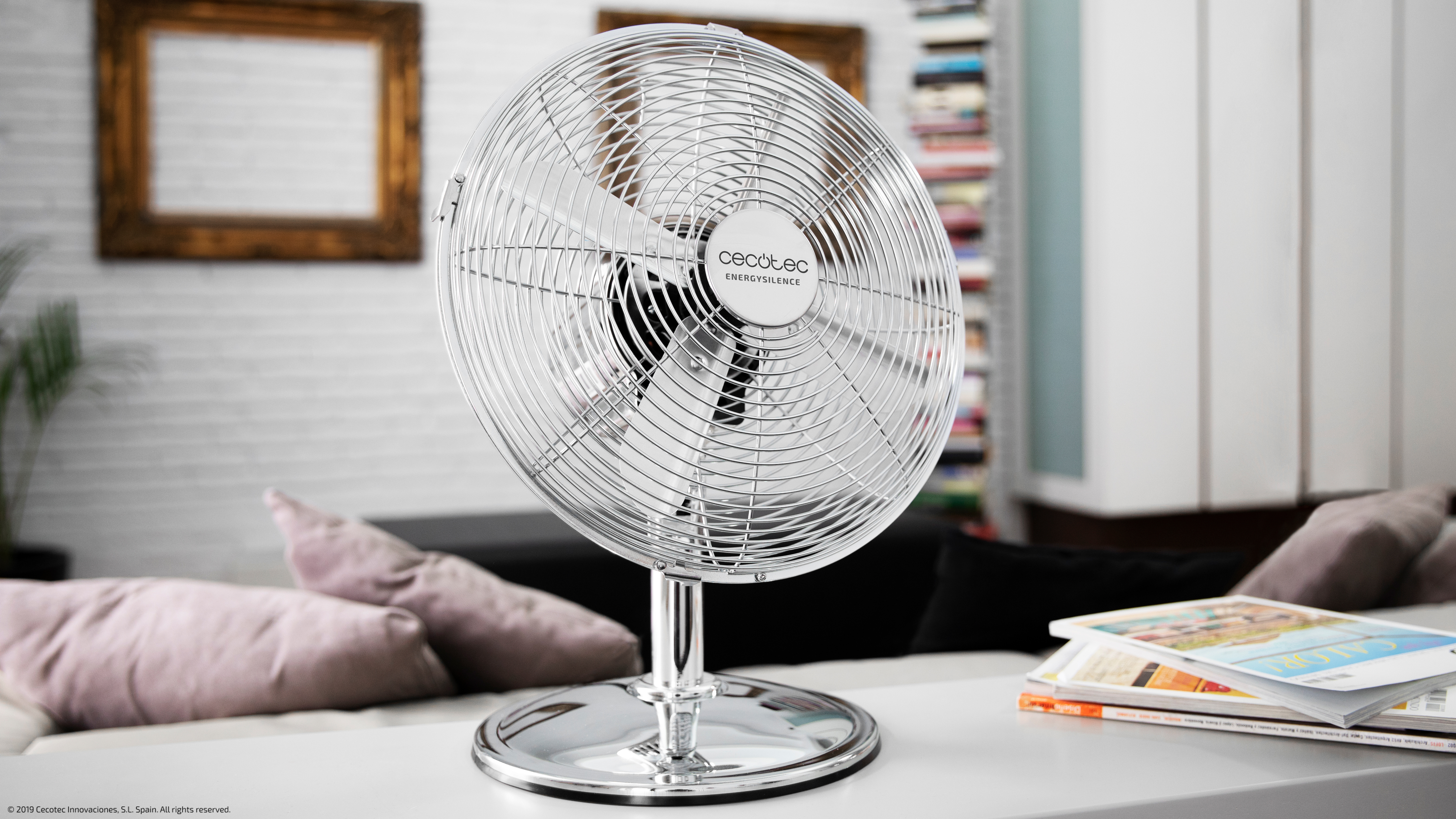 Un ventilador eléctrico plateado sobre un soporte, en una habitación con sofá, revistas y marcos de madera.