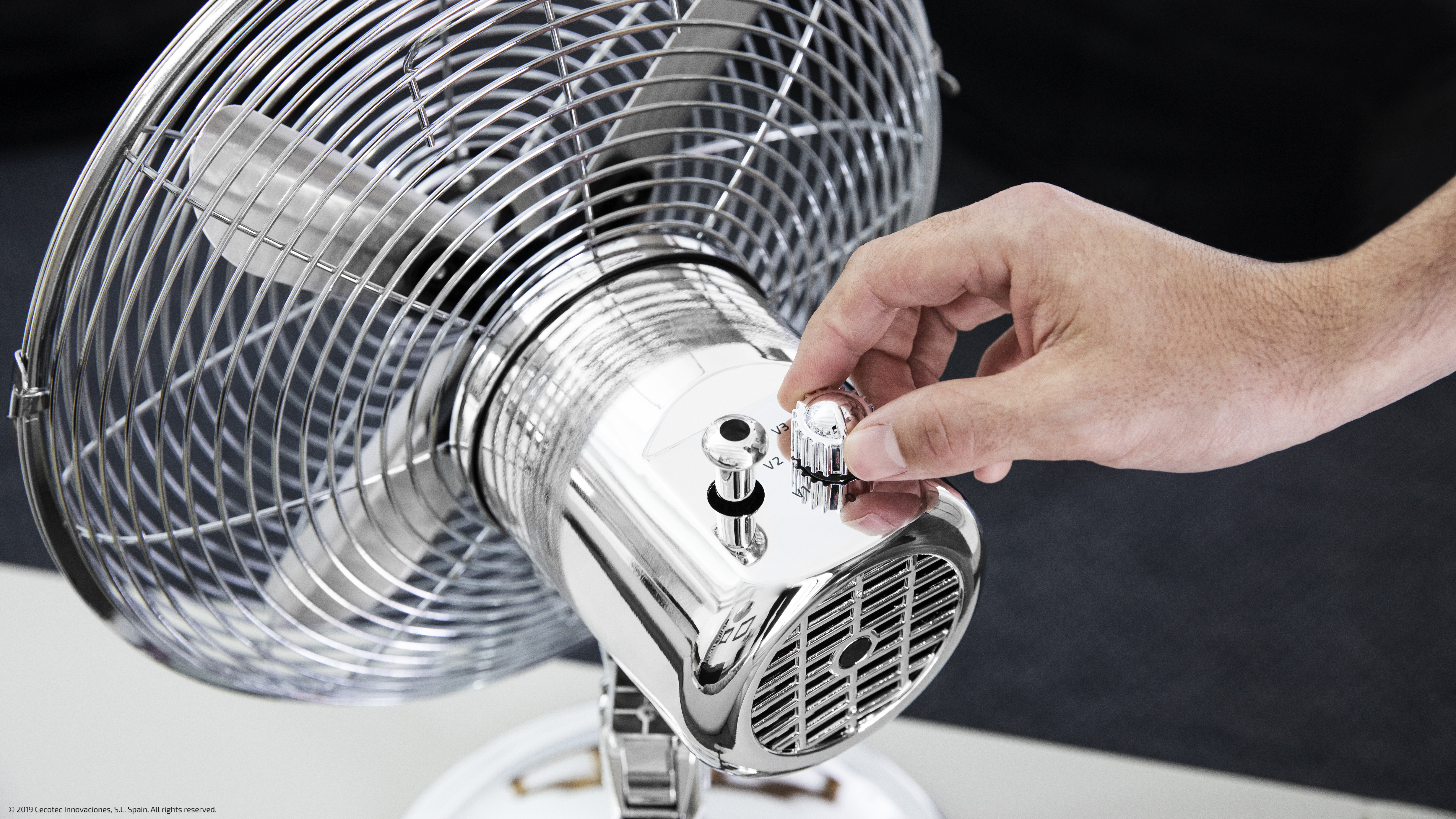 Una mano ajusta la perilla de velocidad de un ventilador retro plateado. El ventilador está en una base blanca.