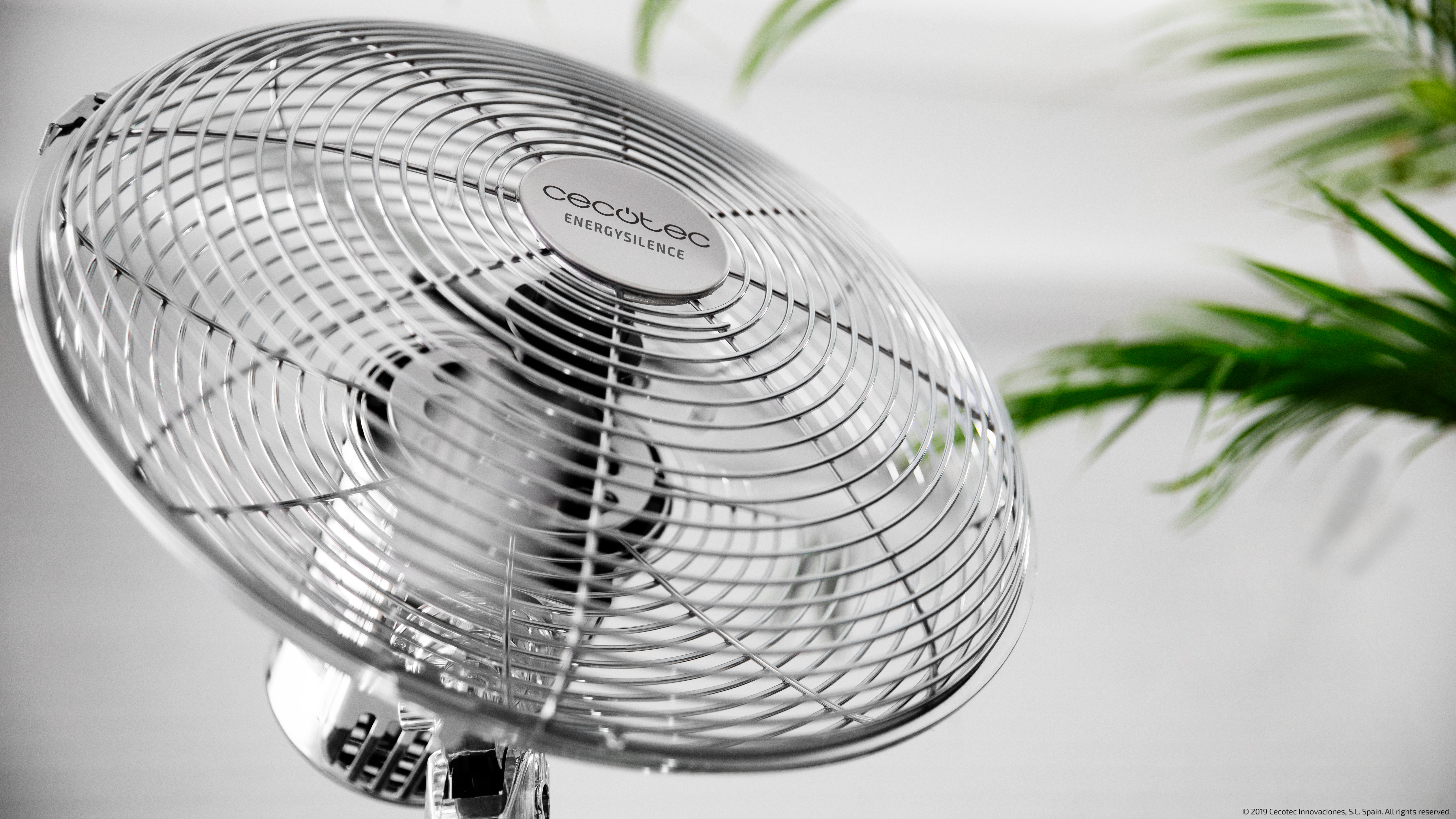 Ventilador plateado con planta verde en bokeh.