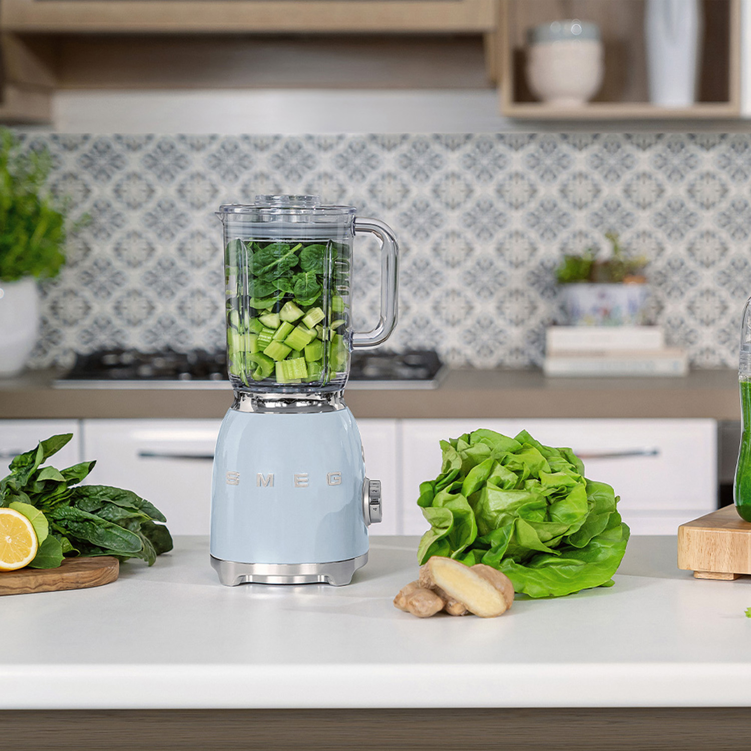 Frullatore azzurro con ingredienti e verdure sul bancone, vicino a una cucina.