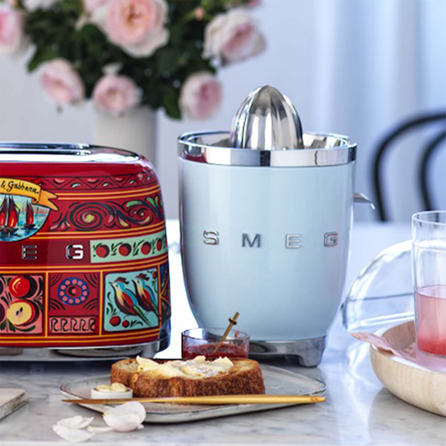 Scena di cucina con spremiagrumi e tostapane Smeg. Toast, marmellata e fiori sono anche sul tavolo.