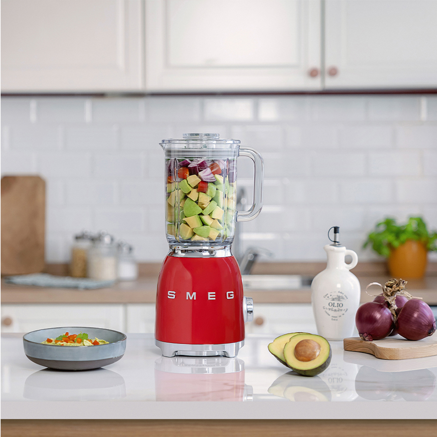 Frullatore Smeg rosso con ingredienti, avocado, cipolle e una ciotola di cibo su un bancone bianco in cucina.