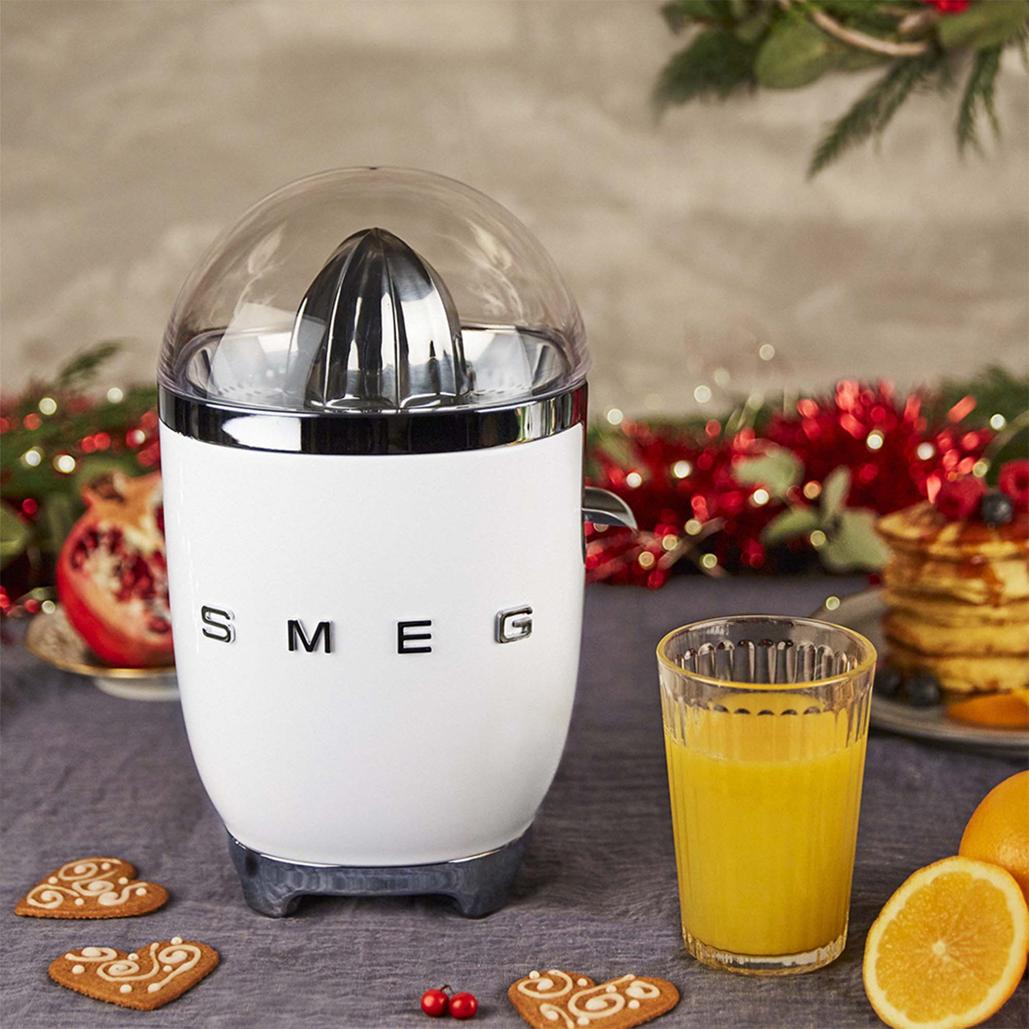 Spremiagrumi SMEG bianco con un bicchiere di succo d'arancia, biscotti e arance su un tavolo. Decorazioni natalizie.