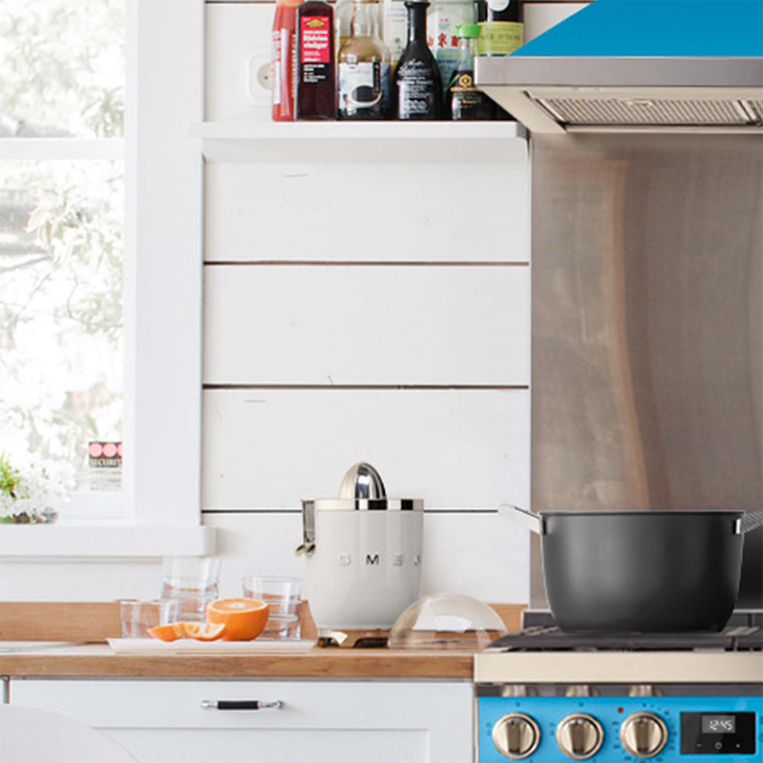 Una cucina con spremiagrumi, arance e una pentola su un fornello blu. Muri bianchi e una finestra sullo sfondo.