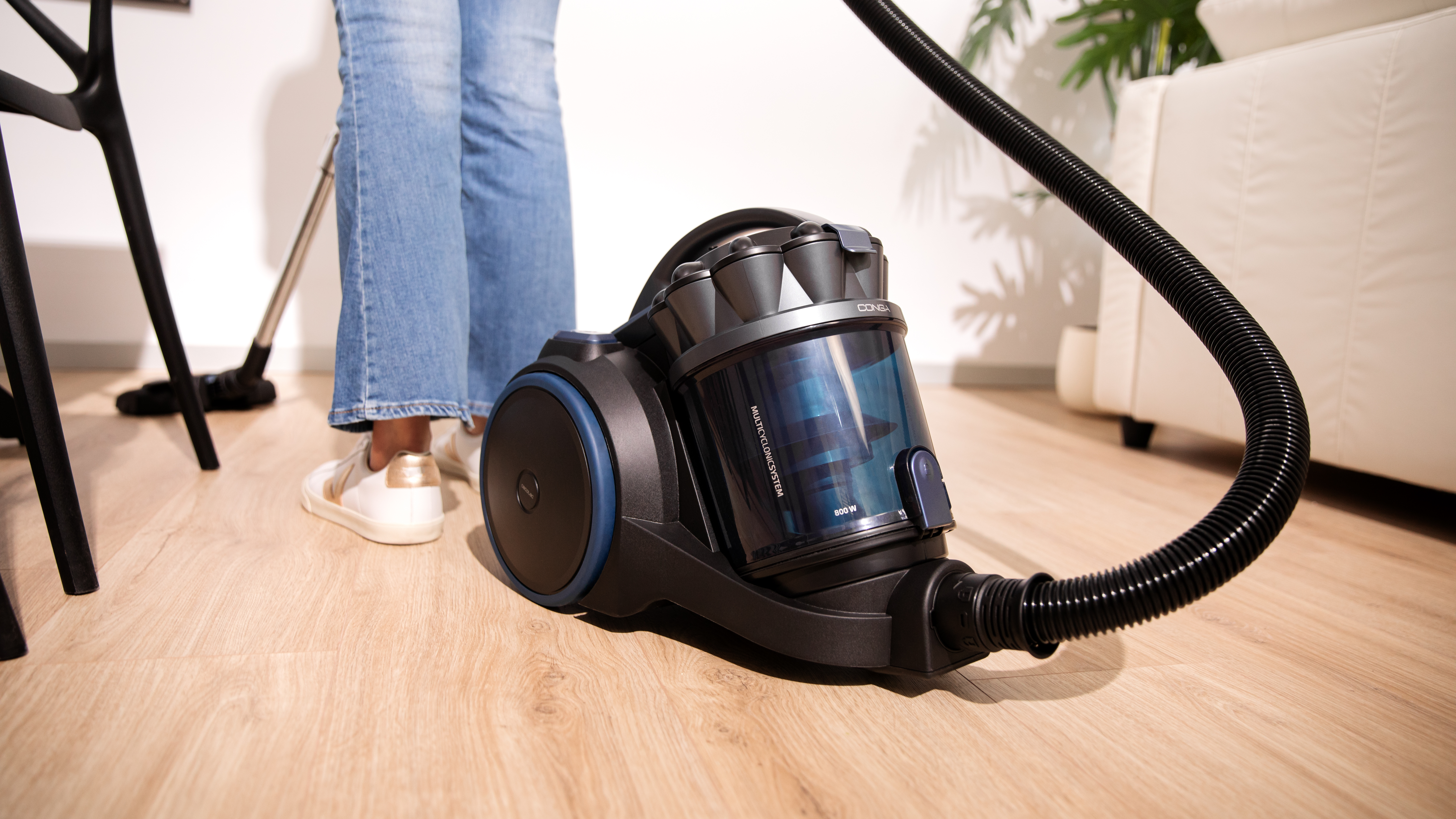 Una persona aspira un suelo de madera con una aspiradora negra. Un sofá y una silla al fondo.