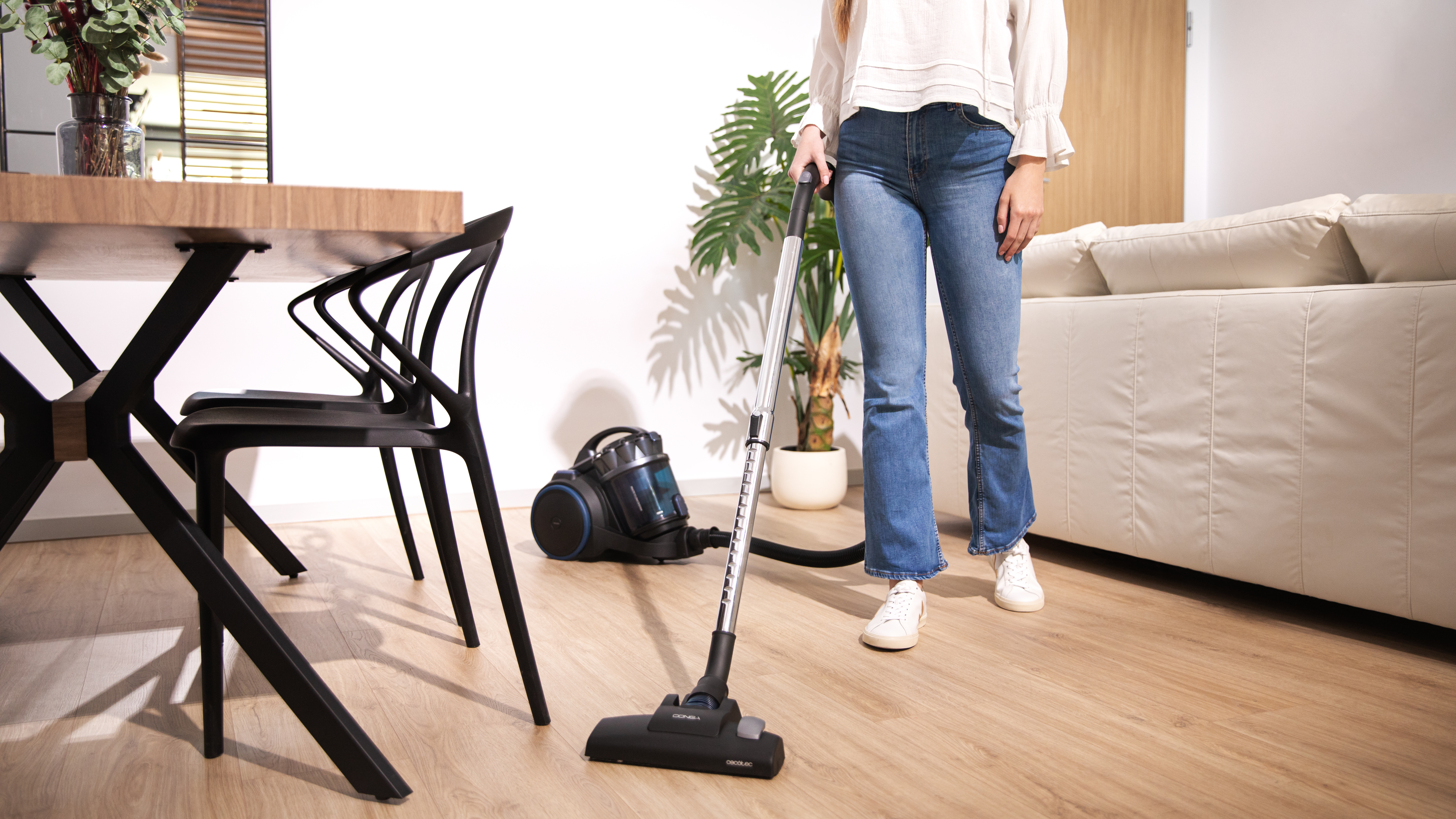 Mujer aspirando una habitación con una aspiradora azul. Suelo de madera y muebles visibles.