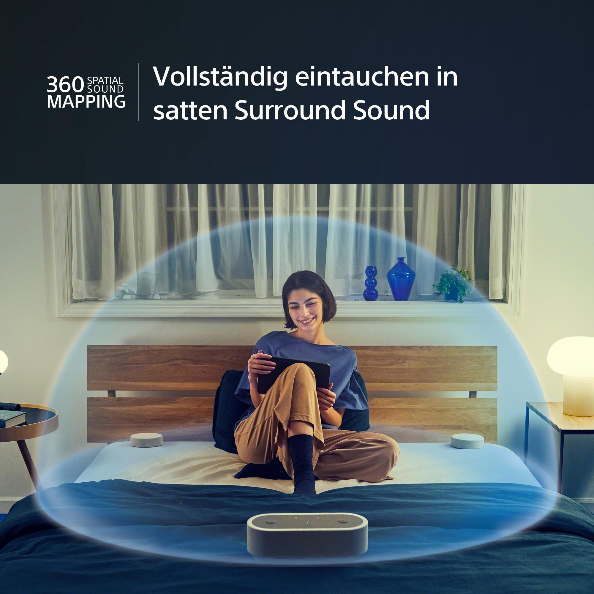 Frau auf dem Bett mit Tablet und Lautsprechern. Umgeben von blauem Licht, weißen Lampen und einem dunklen Schlafzimmer.
