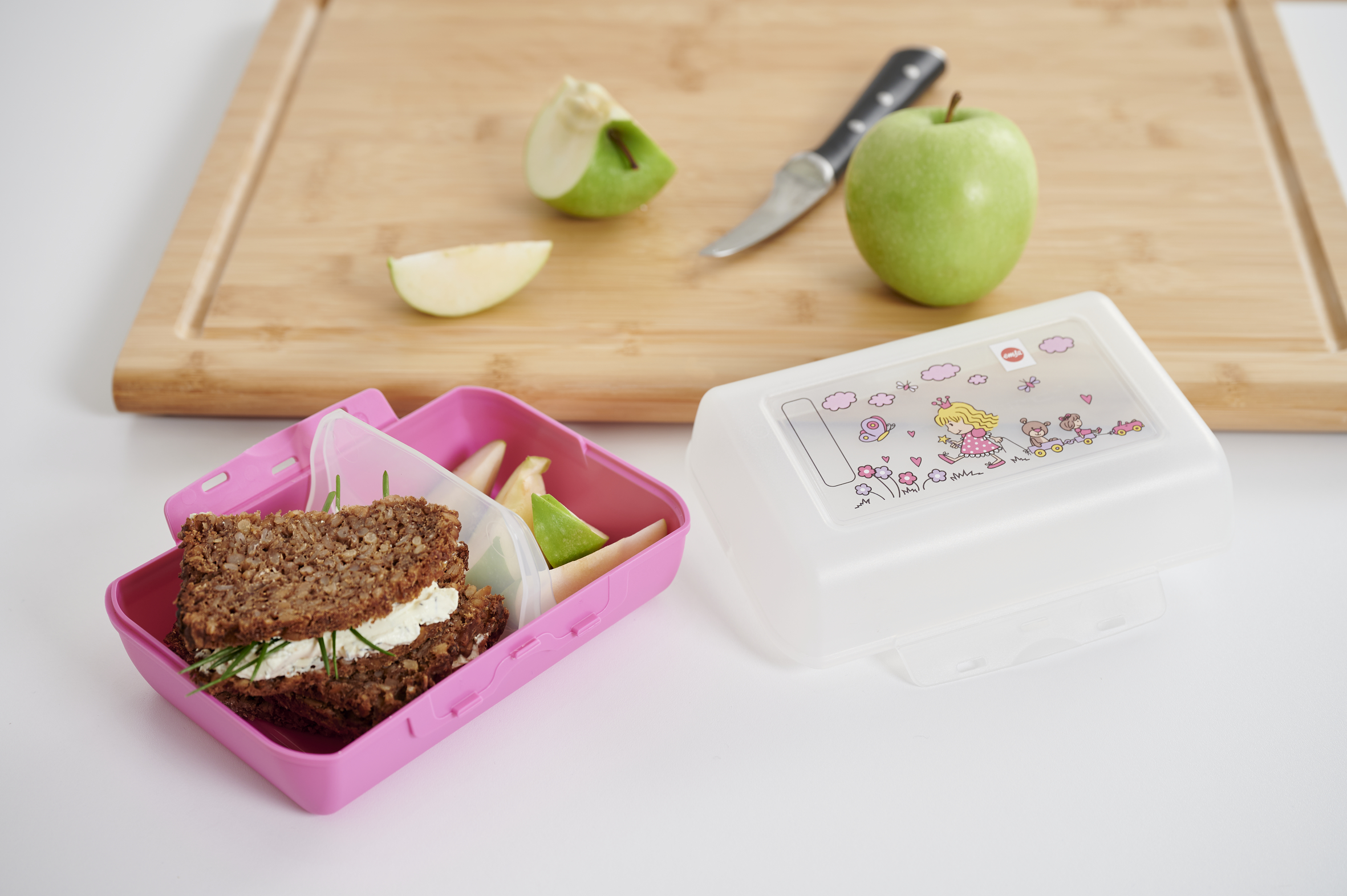 Rosa Brotdose mit Sandwich, Apfelscheiben und einem ganzen Apfel auf einem Holzbrett.