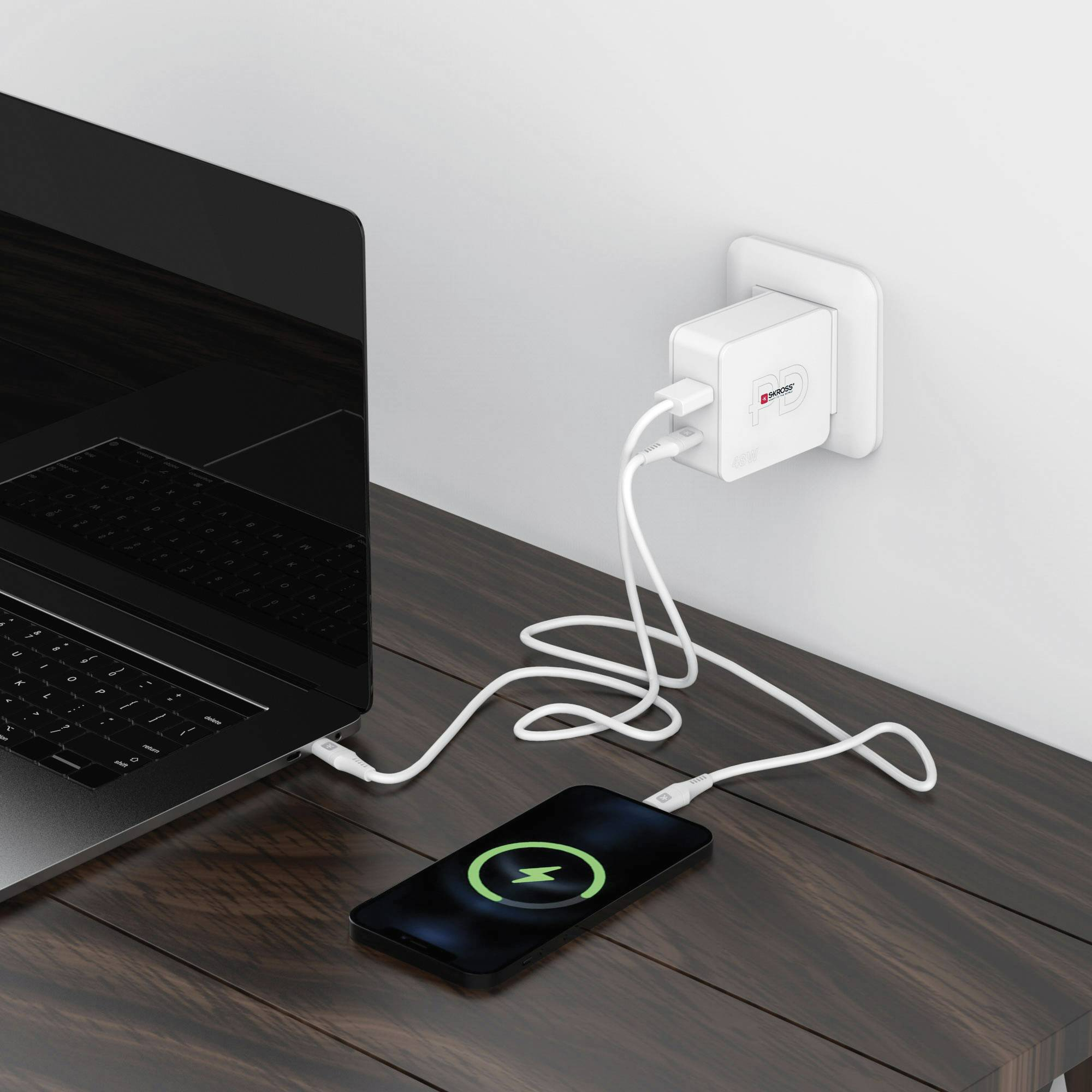 Laptop töltődik egy faasztalon. Egy telefon töltődik mellette. Egy fehér töltő a falon.