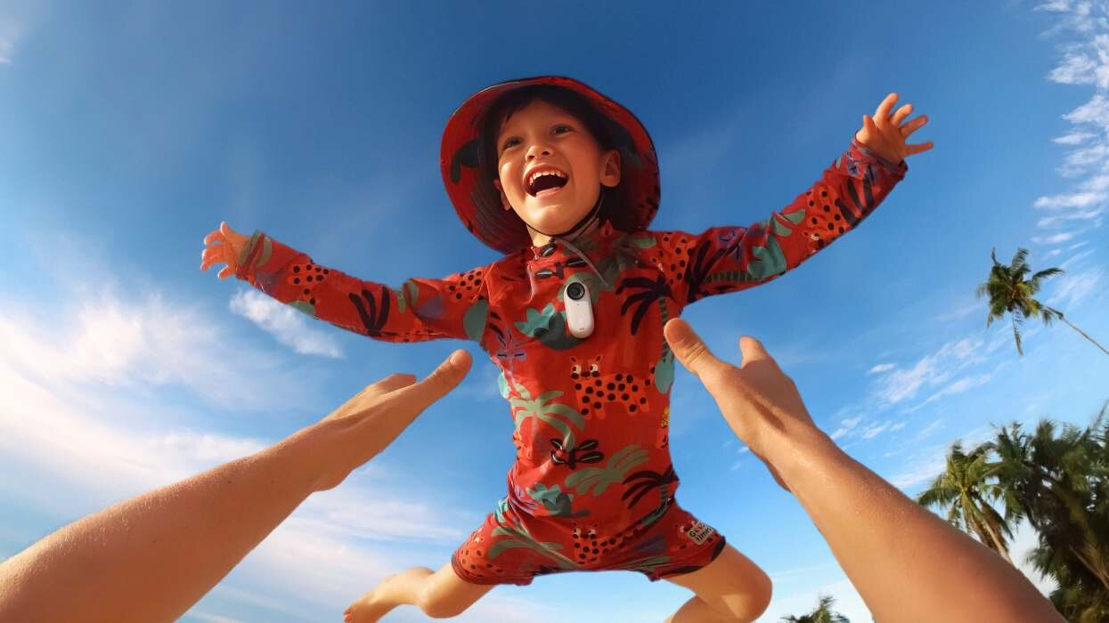 Dziecko w czerwonym stroju kąpielowym leci w powietrzu, trzymane przez dwie ręce, pod błękitnym niebem.