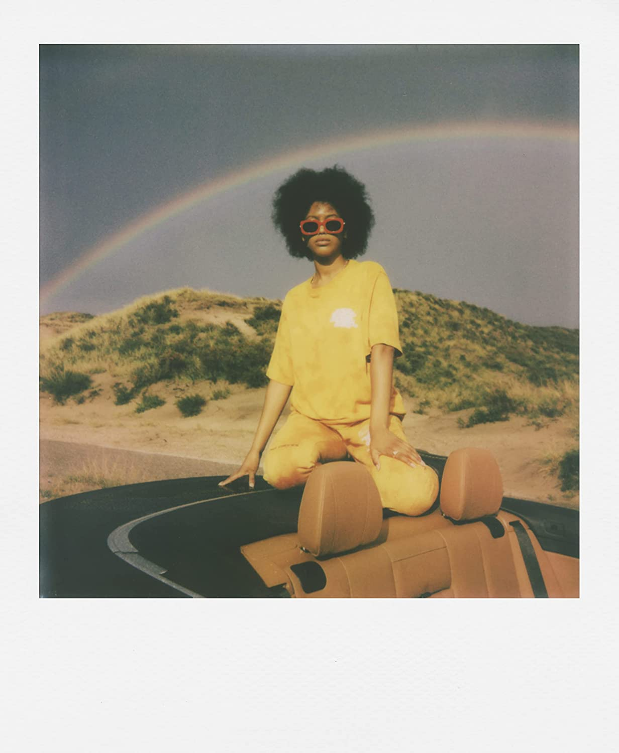 Sarı kıyafetli bir kadın, arka planda gökkuşağı ve polaroid çerçevesi olan bir araba üzerinde oturuyor.