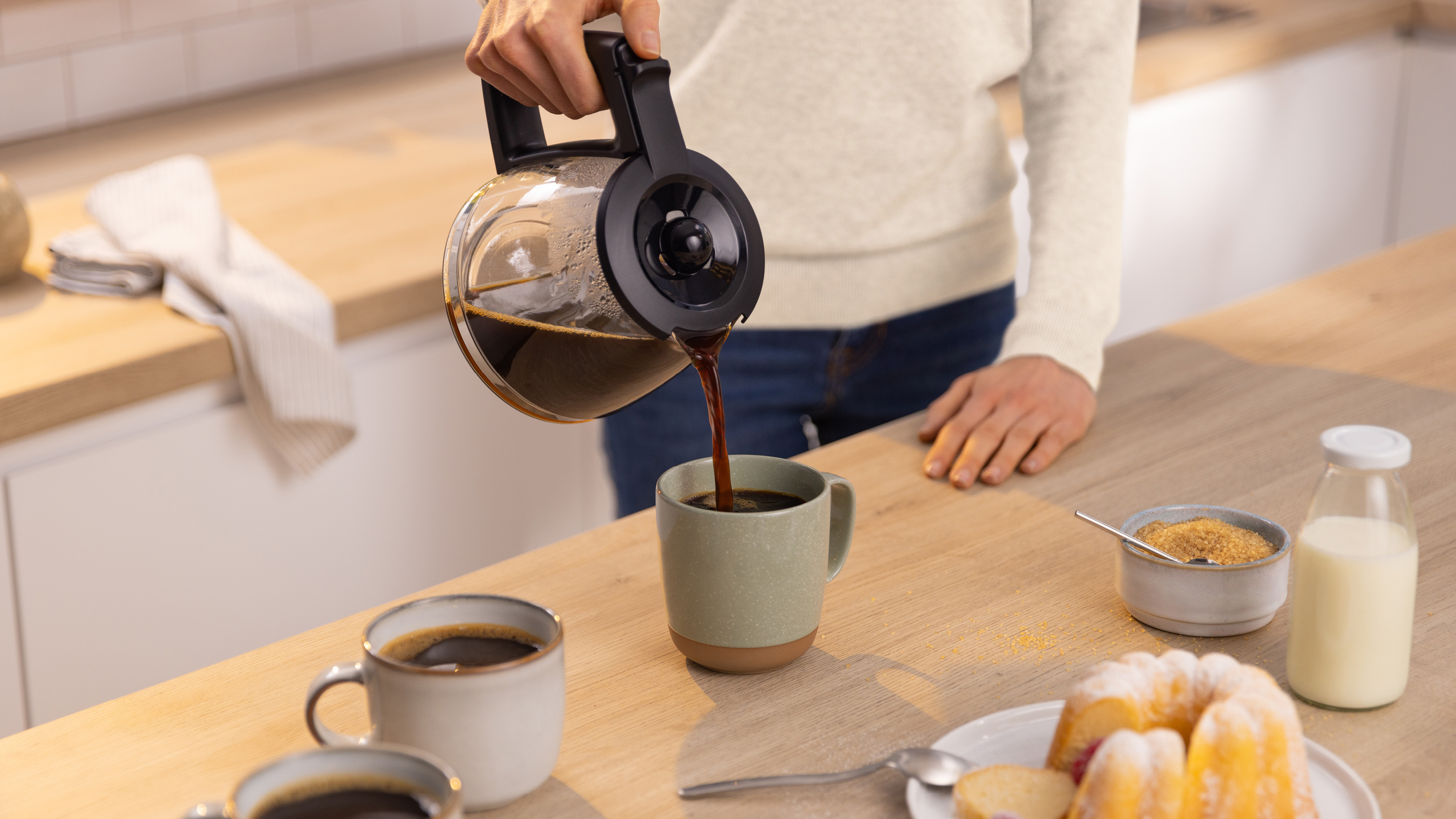Eine Person gießt Kaffee aus einer Karaffe in eine Tasse, mit Kaffee, Milch und Kuchen auf dem Tisch.