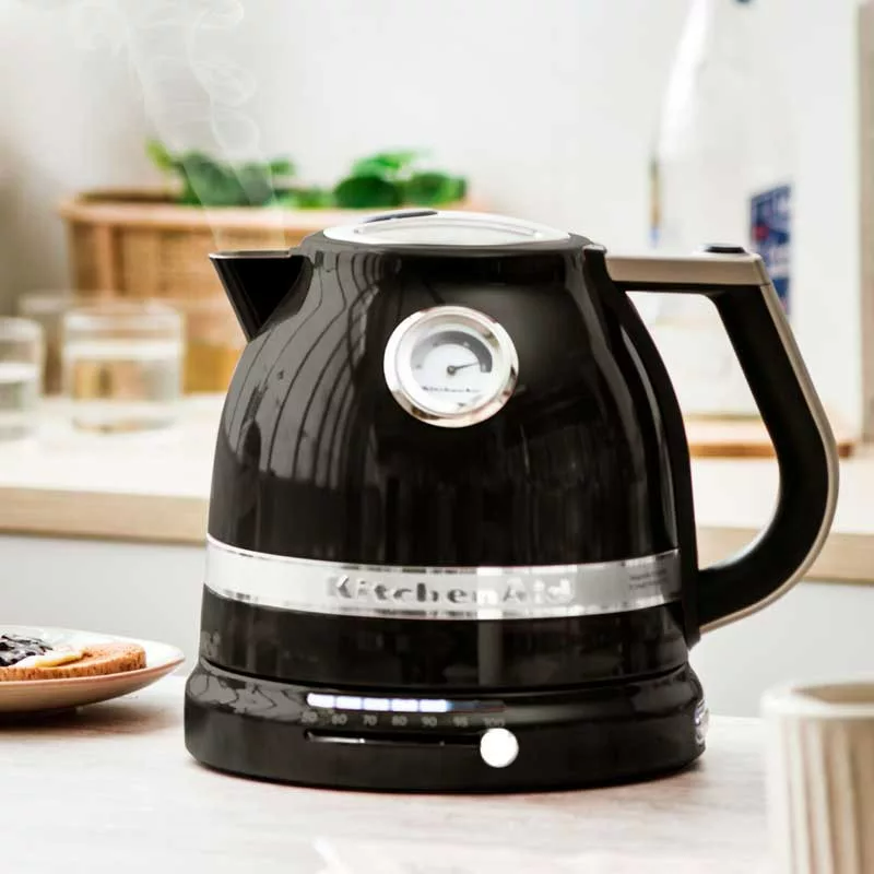 Fekete KitchenAid vízforraló gőzzel, számlappal és fogantyúval. Pirítós lekvárral elöl, fa felület. Háttér: üveg, zacskó és zöldségek.