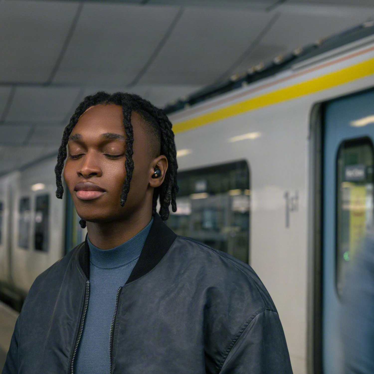 Een man met dreadlocks draagt een blauwe trui en zwarte oordopjes in een metrostation, ogen gesloten.