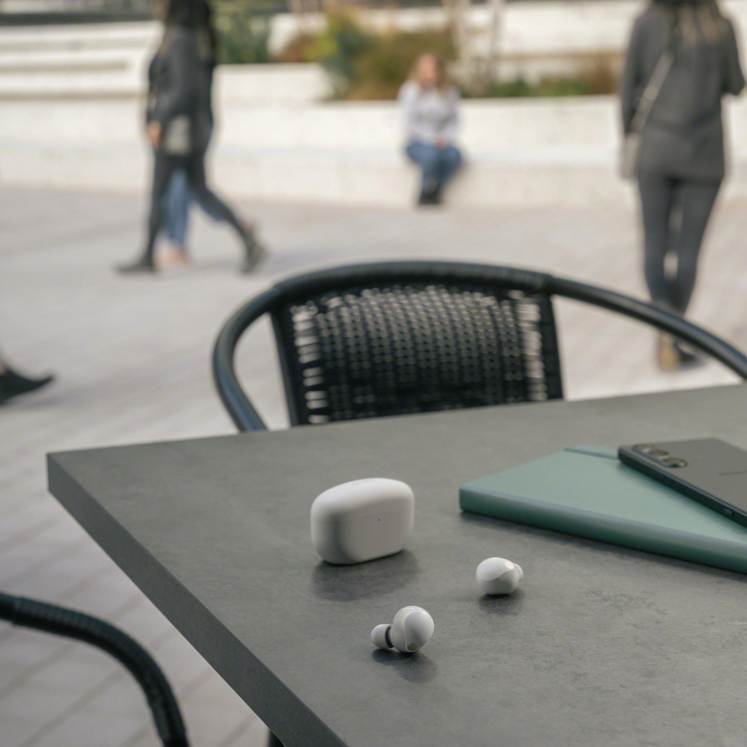 Oordopjes en hoesje op een tafel met telefoon en notitieboekje; mensen vaag op de achtergrond.